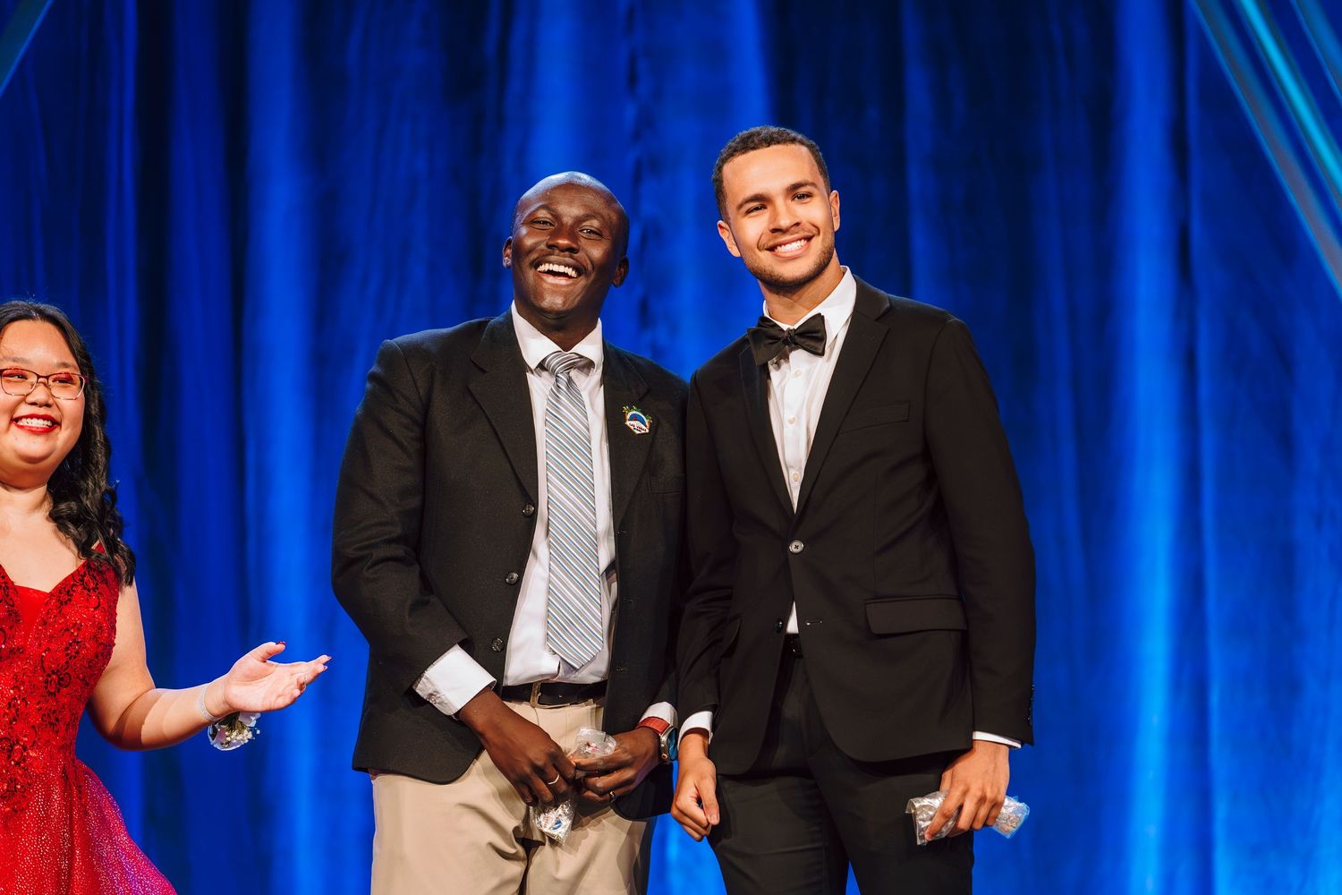 Two men in formal attire smiling on stage with blue curtain backdrop.