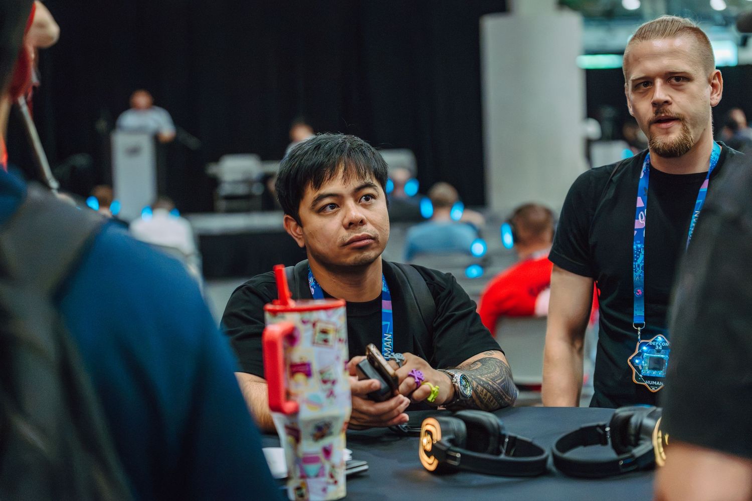 Two men at a gaming convention table, one holding a controller, with headphones and drinks nearby.