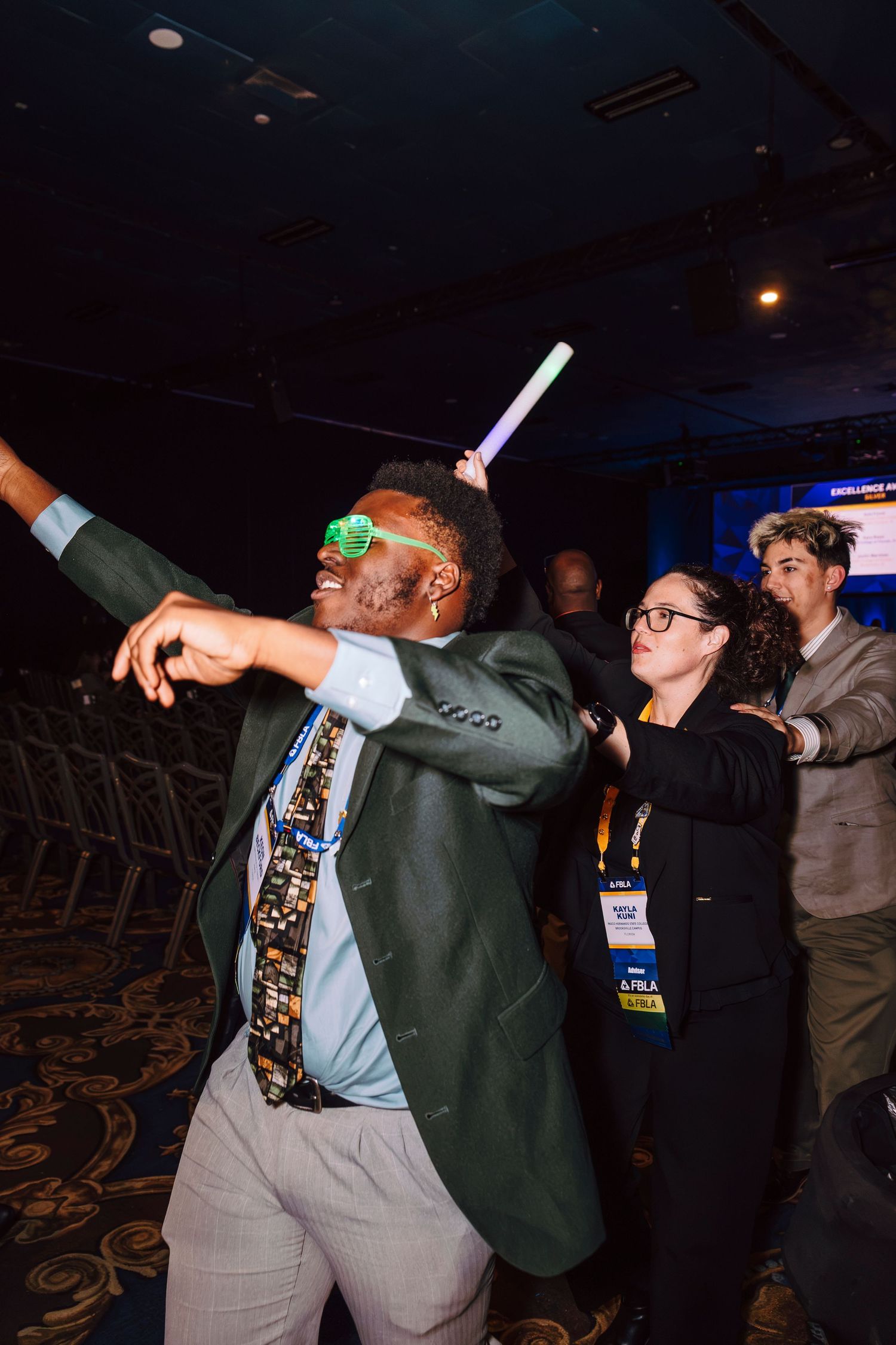 Man in green blazer and novelty tie dances with glow stick at a conference event party.