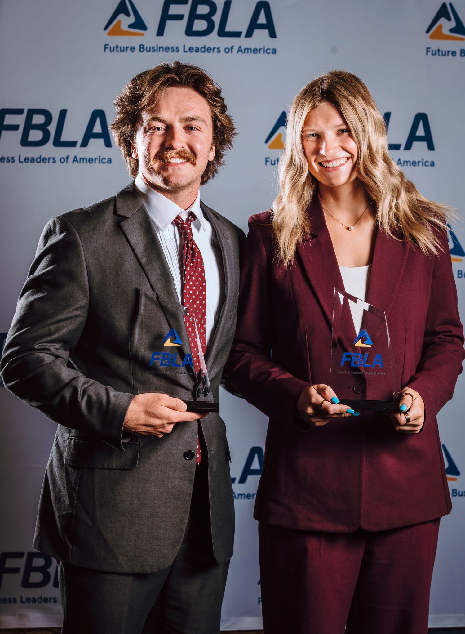 Two FBLA award winners in maroon suits proudly holding crystal trophies at a Future Business Leaders of America event.