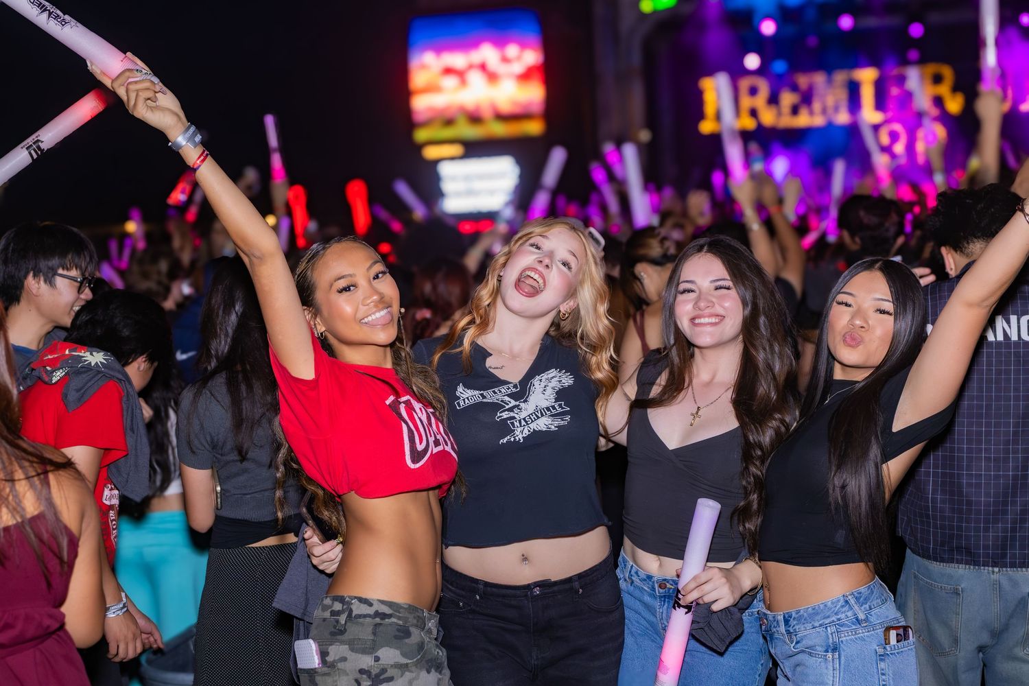 Group of young women dancing and celebrating at a concert event with colorful glow sticks and crowd.