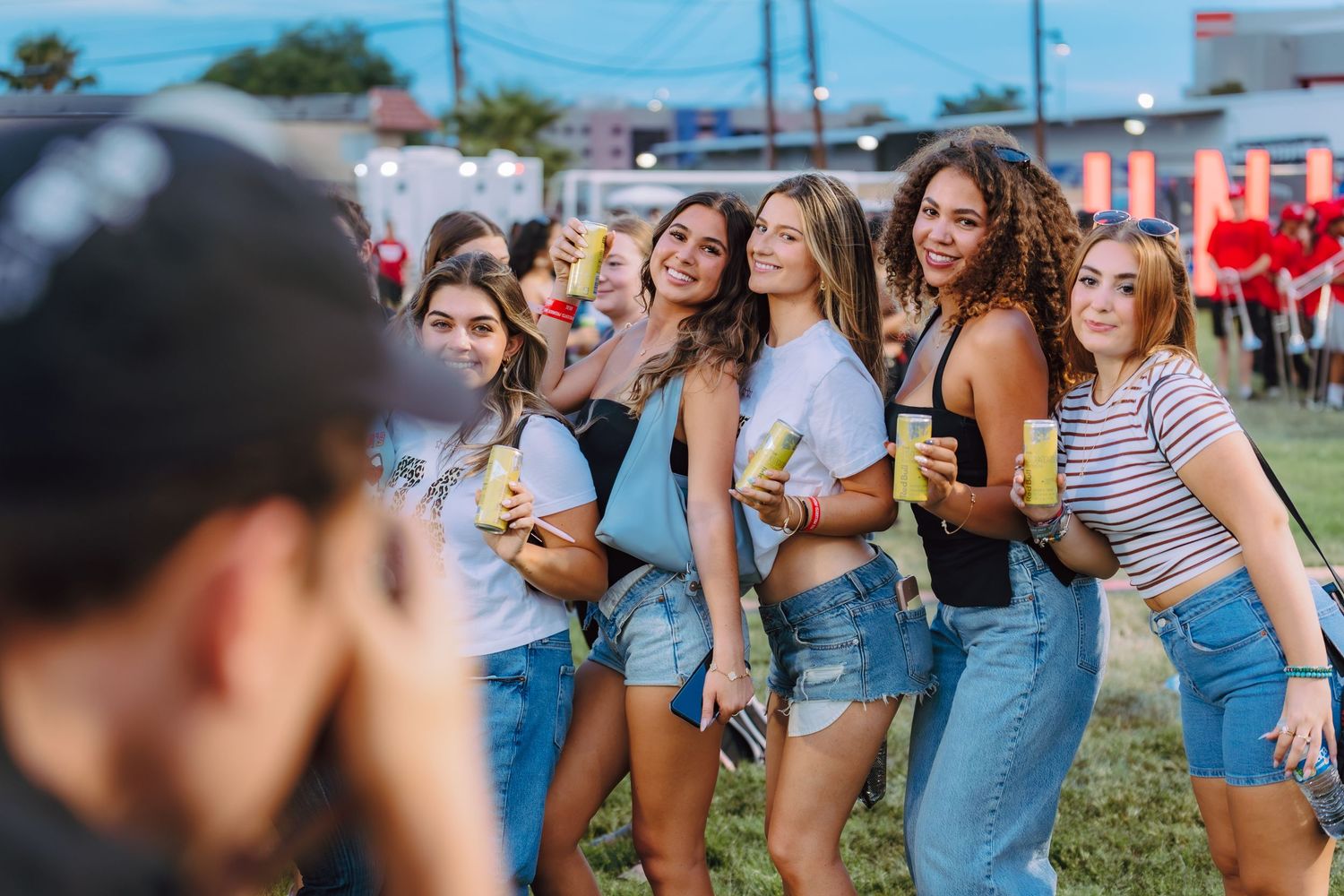 Group of young women posing together at an outdoor festival, holding drinks and smiling for a photo.