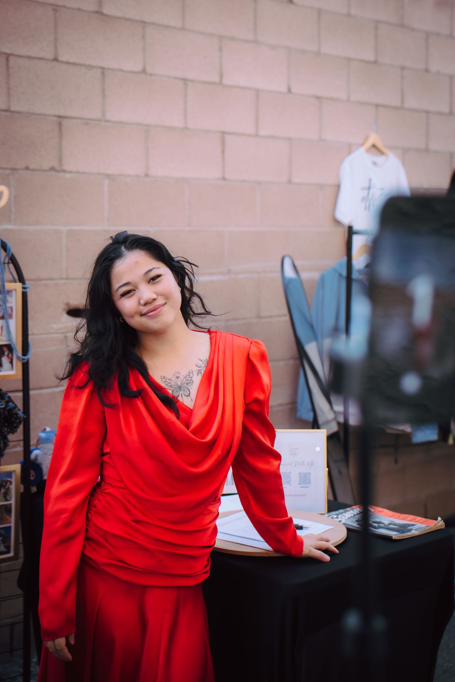 Woman in red outfit smiling at vendor booth with white t-shirt display and brick wall background.