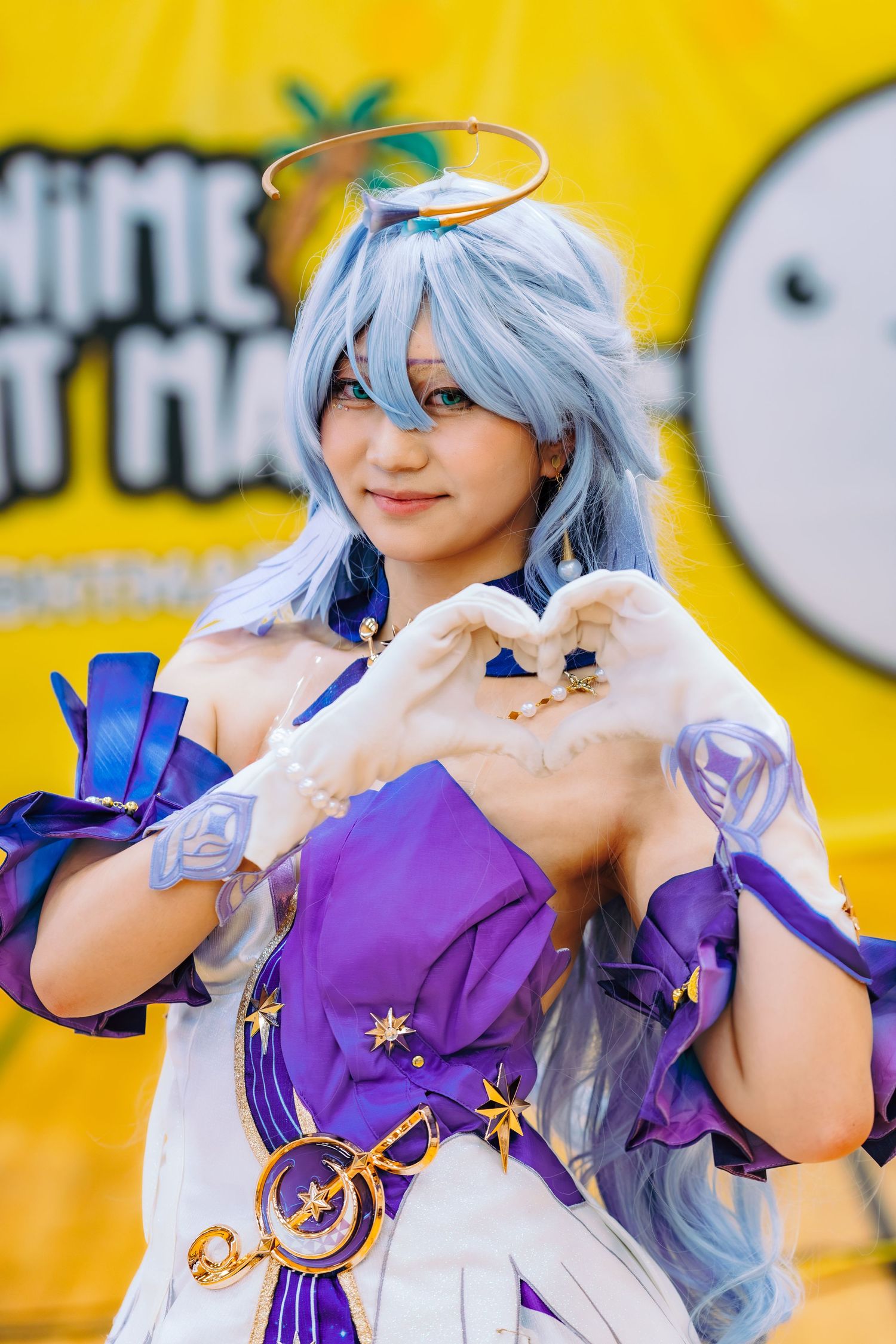 Cosplayer in blue wig and purple costume makes heart shape with hands at anime convention.
