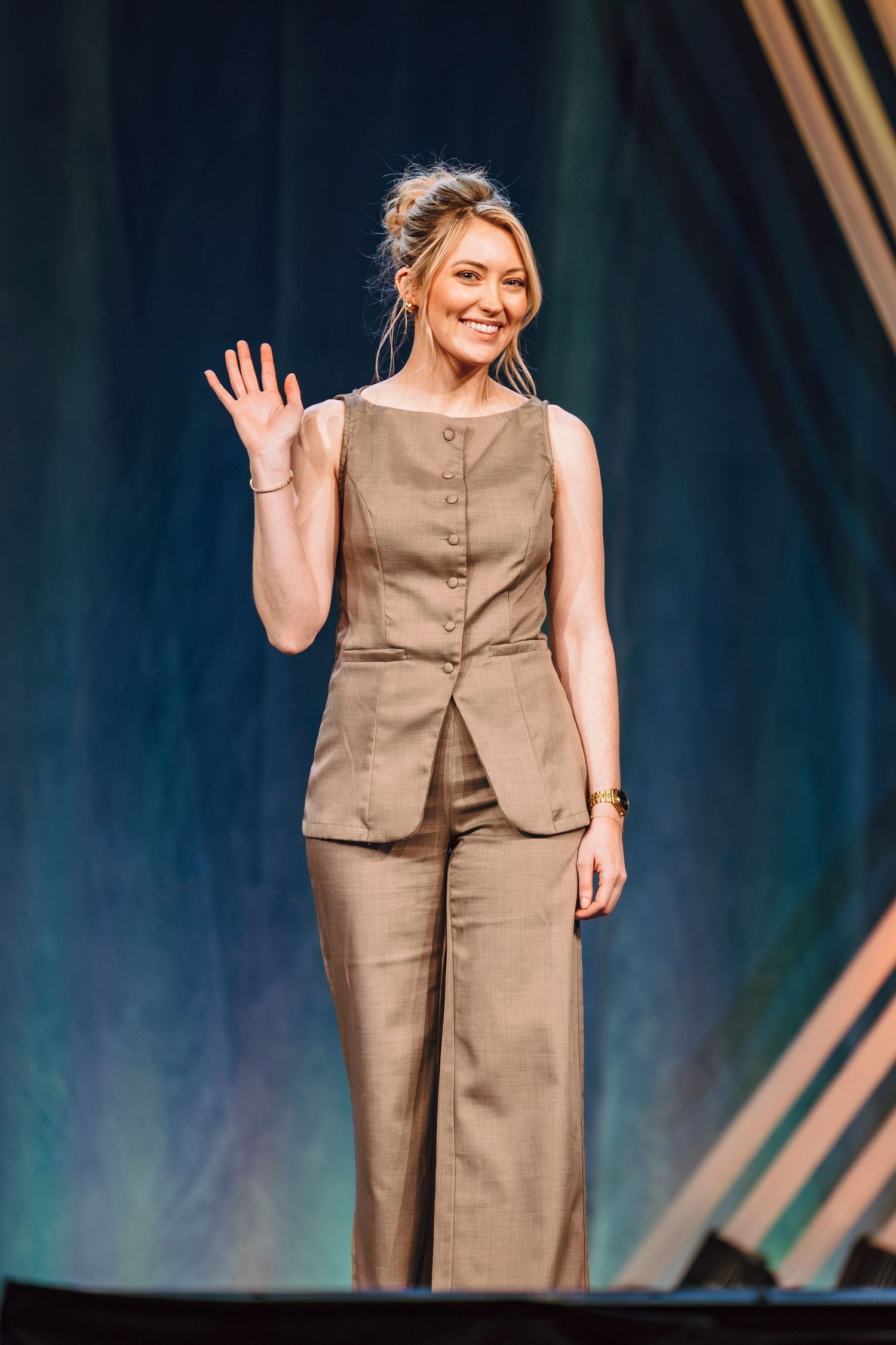 Woman in beige pantsuit waving and smiling on stage at a formal event.