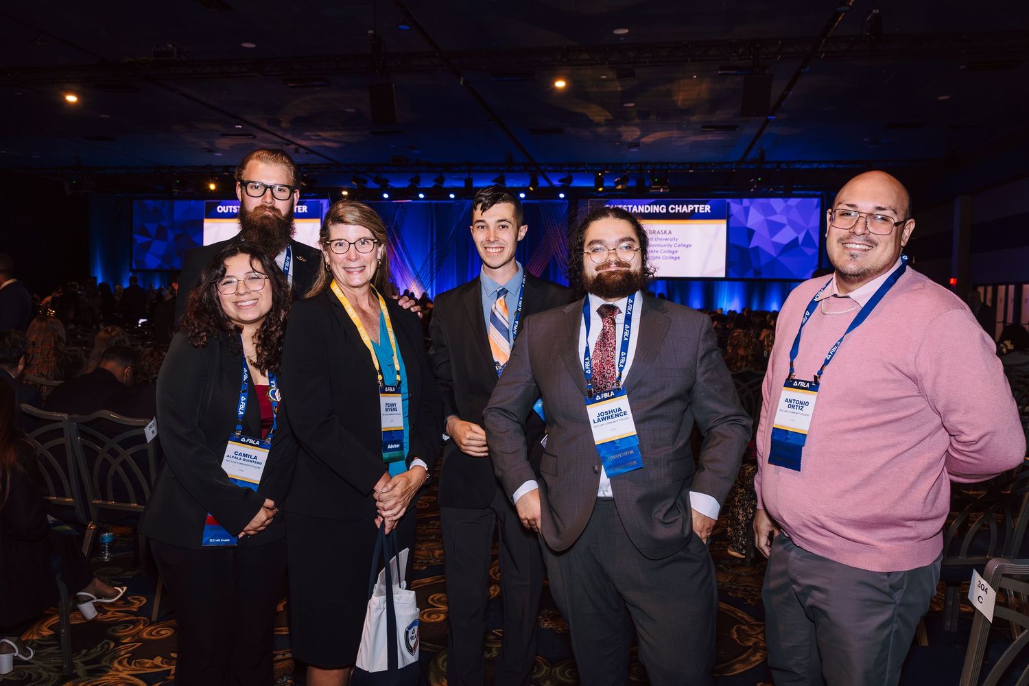 Six professionals wearing conference badges pose together smiling at a formal networking event with blue stage lighting.