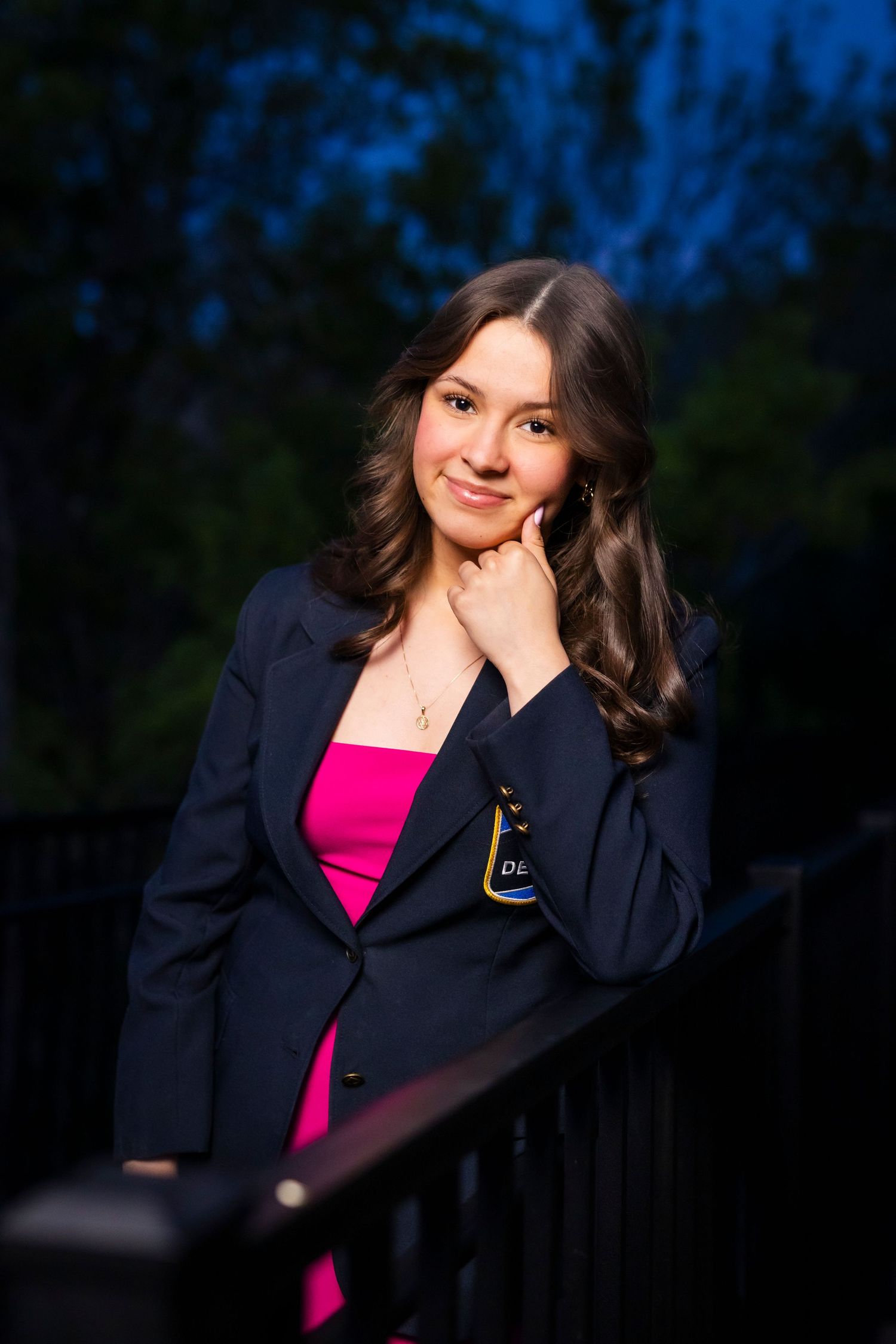 Young woman in navy blazer and pink top poses gracefully on a deck at dusk with trees in background.