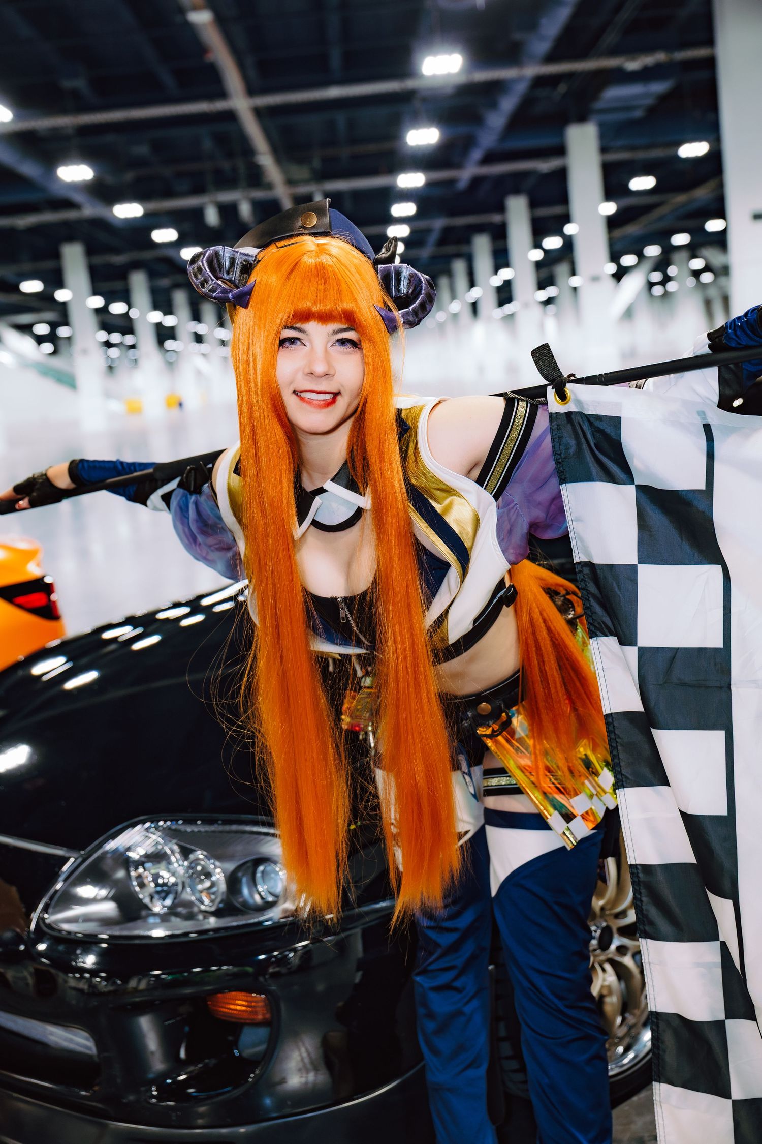Woman in orange wig and anime cosplay costume poses with checkered flag beside a black car at a convention.