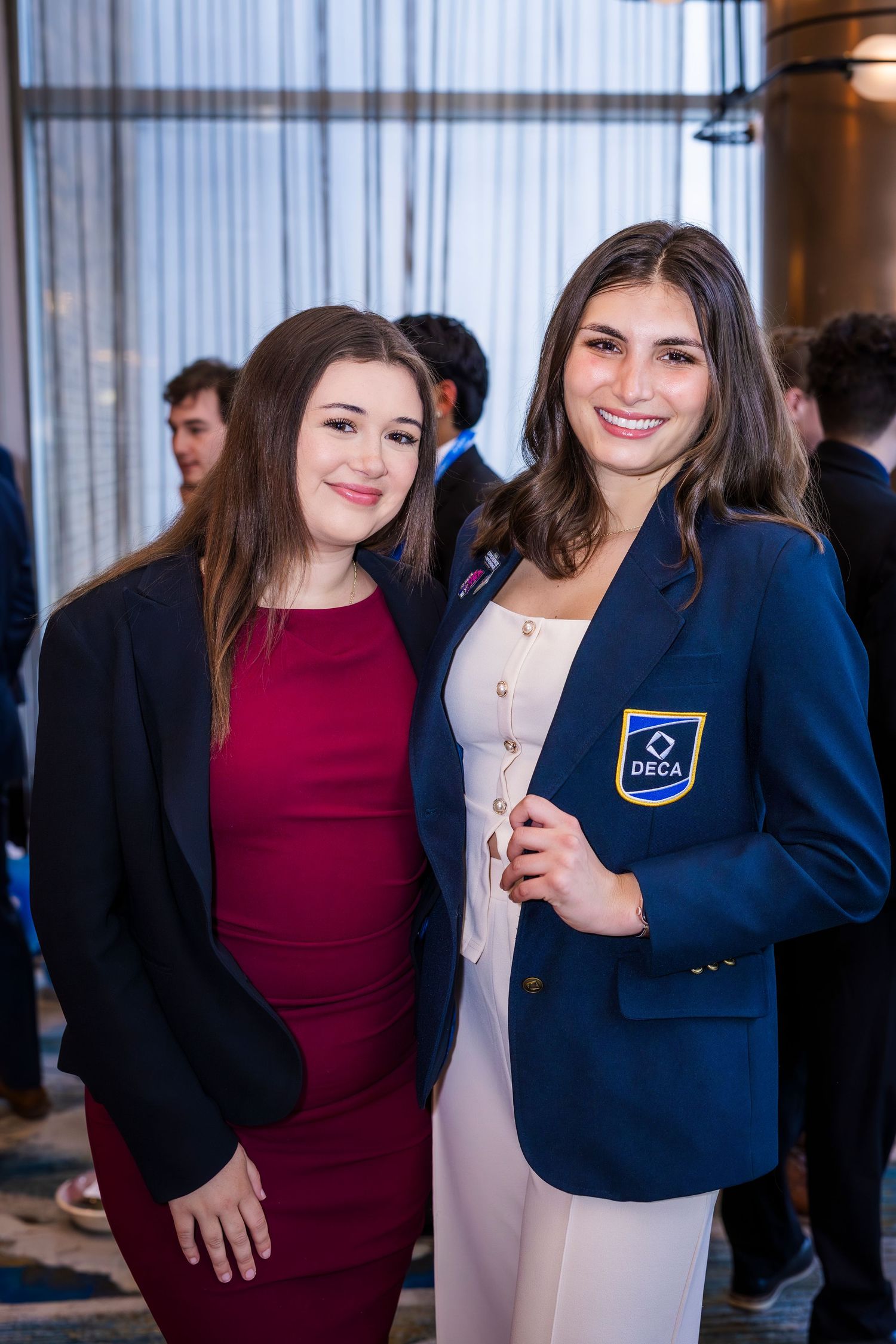 Two young women posing together, one wearing an official navy DECA blazer at an event.