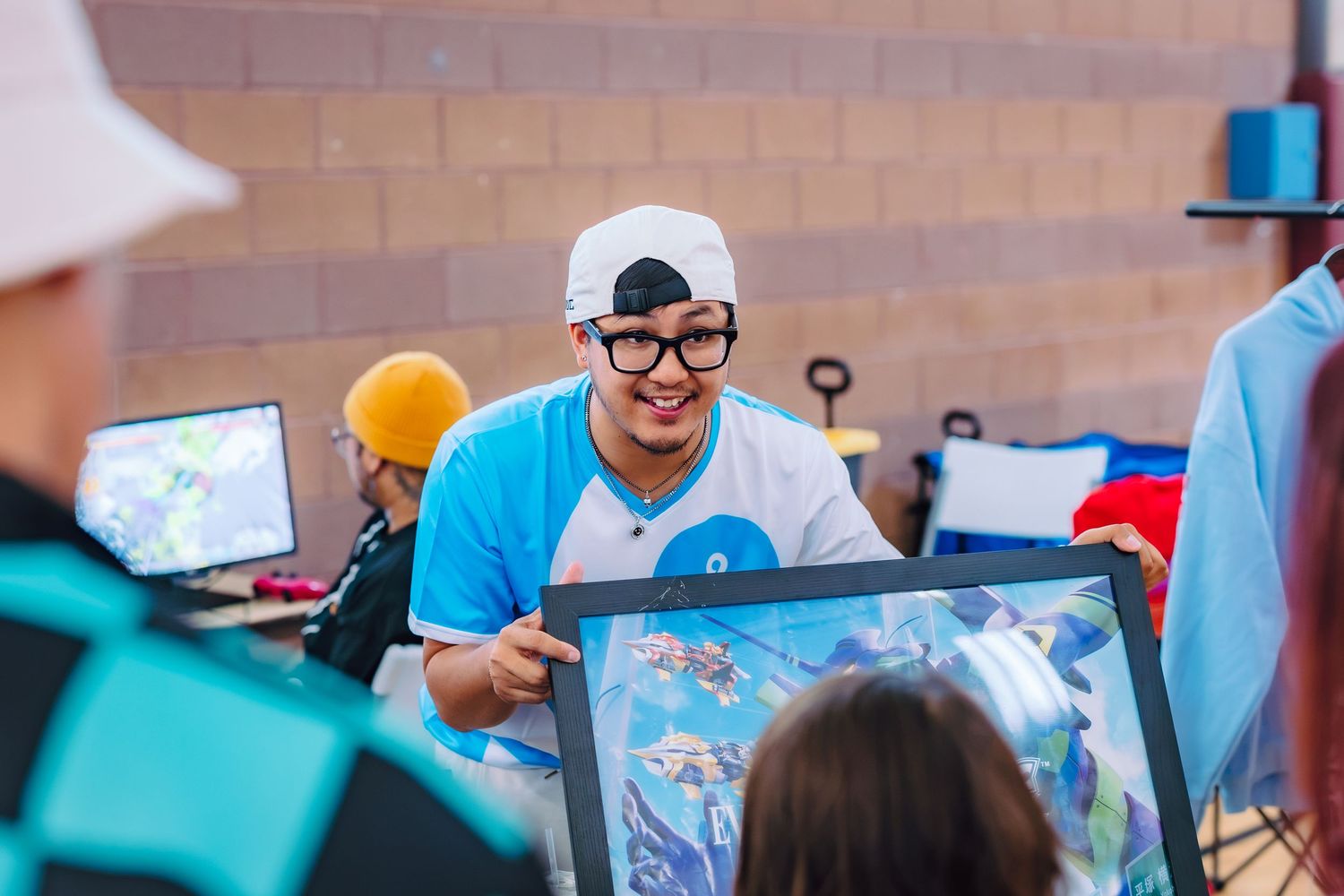 Smiling man in white cap and glasses holds framed artwork at an outdoor gaming event.