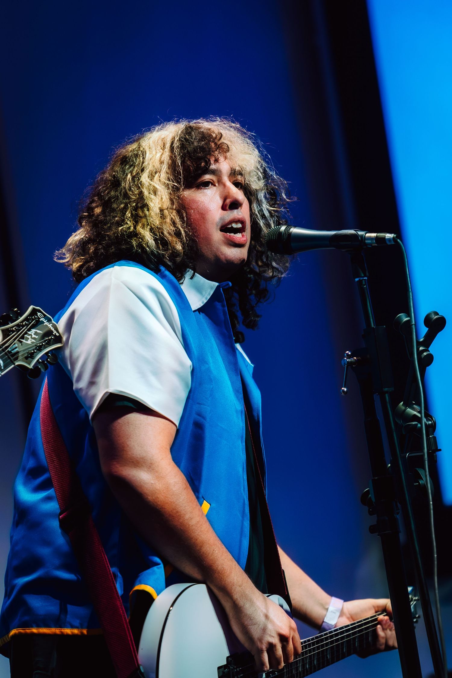 Young curly-haired musician singing into microphone while playing guitar on stage with blue lighting.