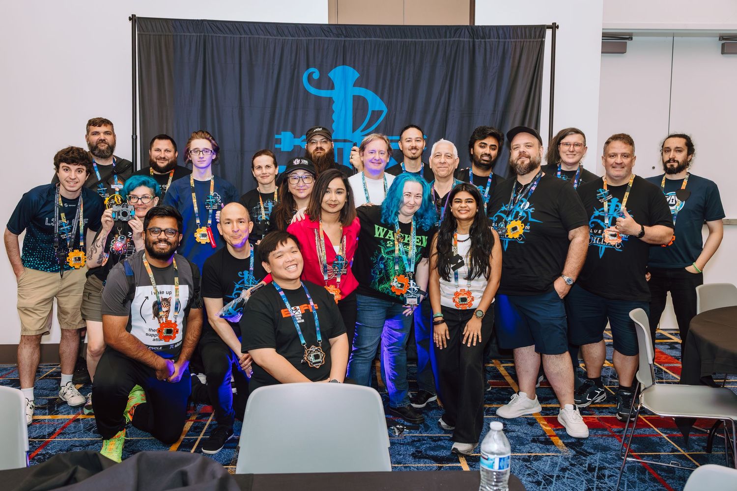 Large group of smiling people posing together at a gaming convention event with a blue logo banner backdrop.