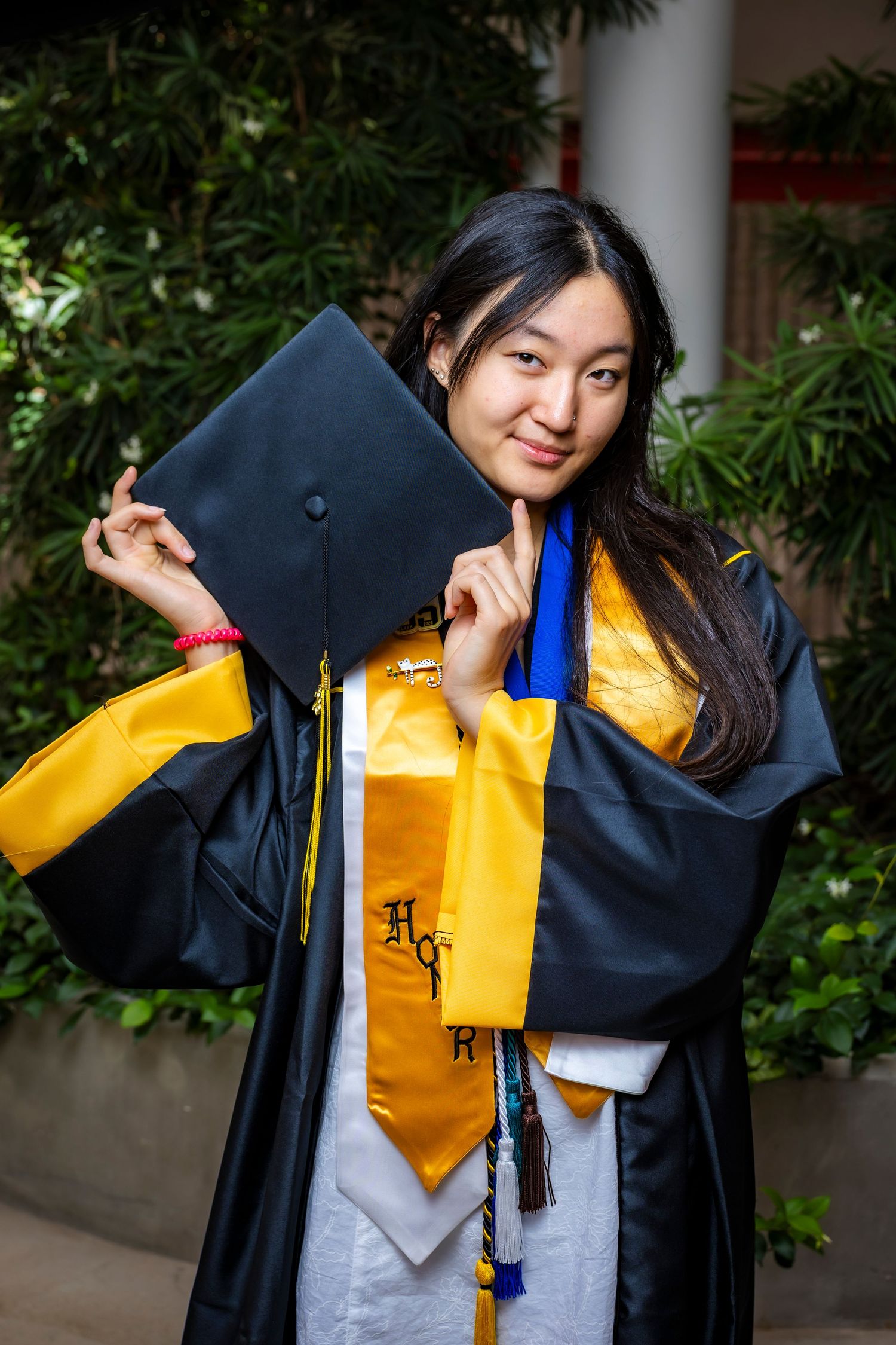 Smiling graduate poses in black and gold cap and gown holding mortarboard with blue and gold honor cords.