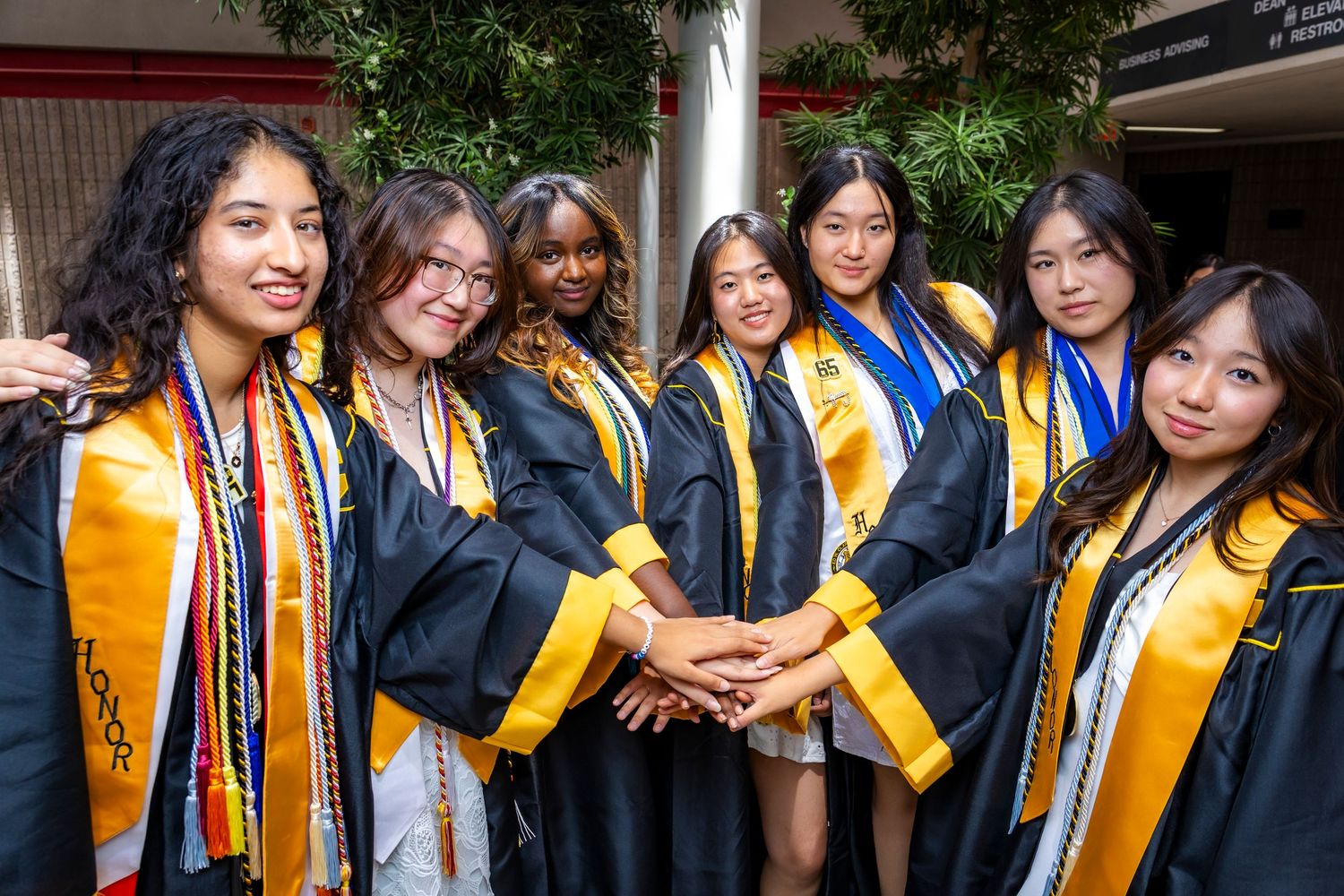 Seven female graduates in black and gold gowns smiling and stacking hands together at graduation ceremony.