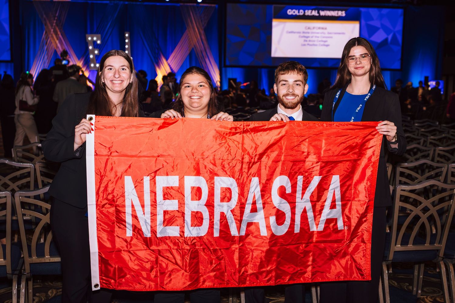 Four students proudly hold a red Nebraska flag at an awards ceremony in a decorated conference hall.