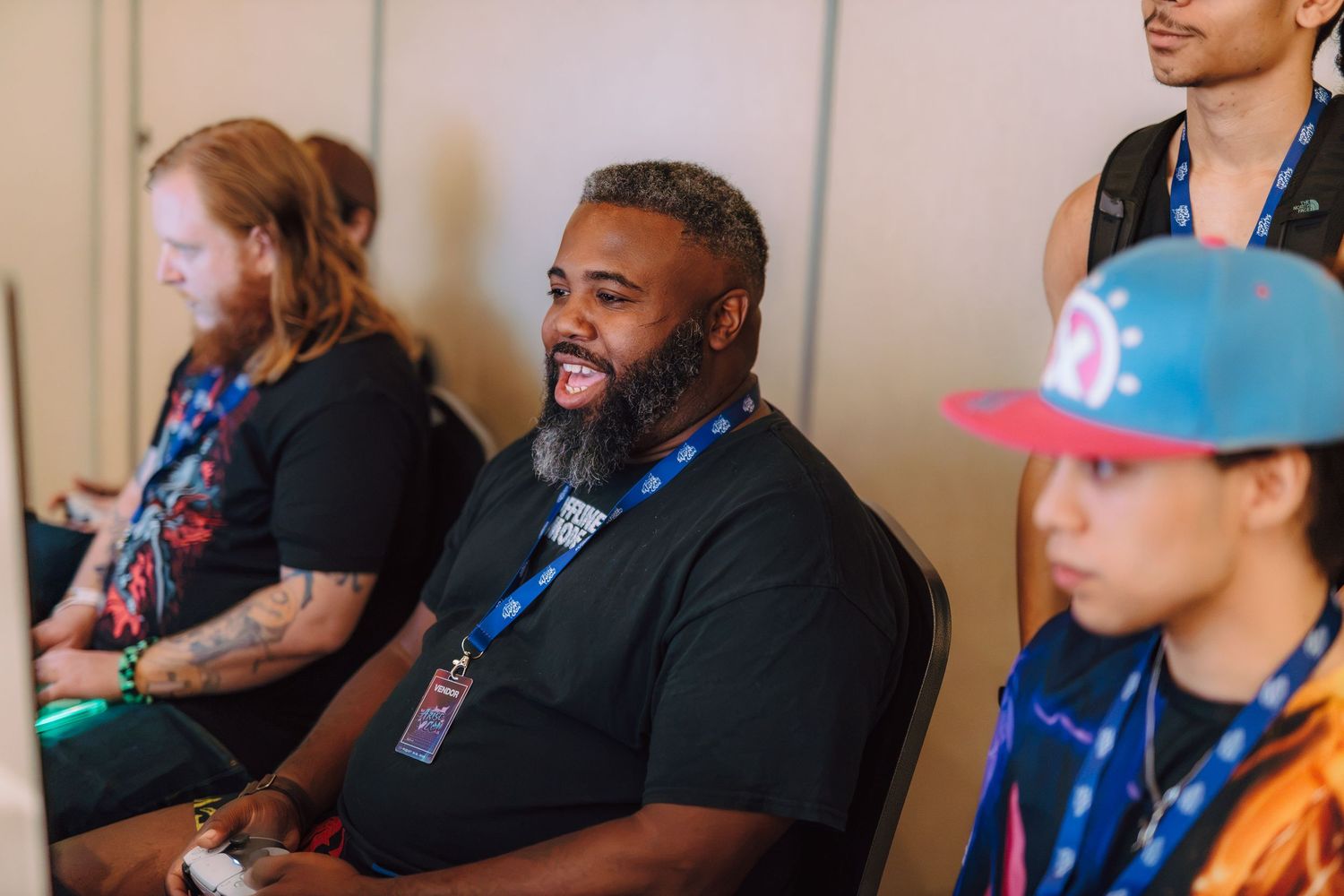 Three people sitting at a gaming event, smiling and laughing, wearing lanyards and colorful caps.