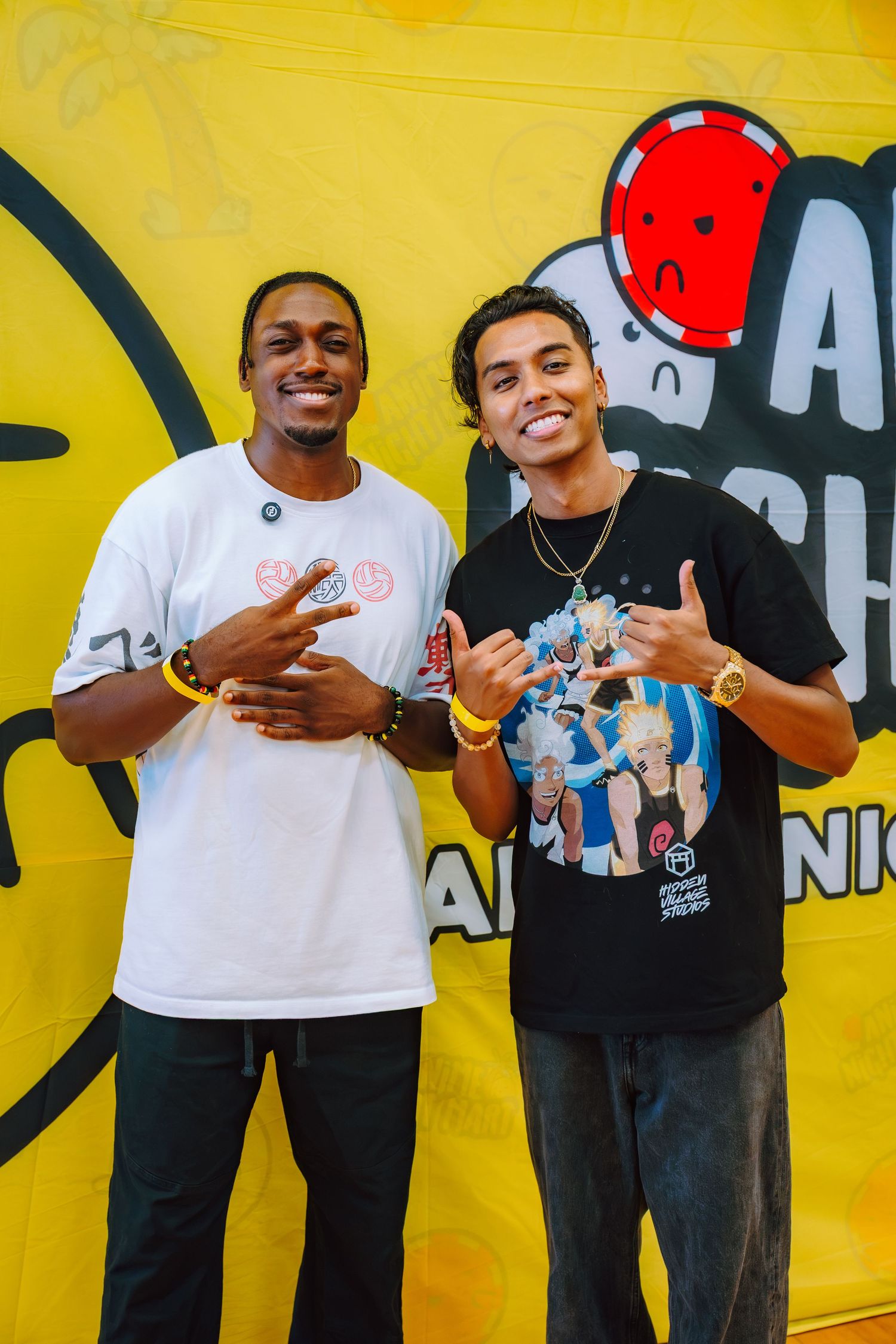 Two young men posing together in front of a yellow banner, wearing graphic tees and gold jewelry.
