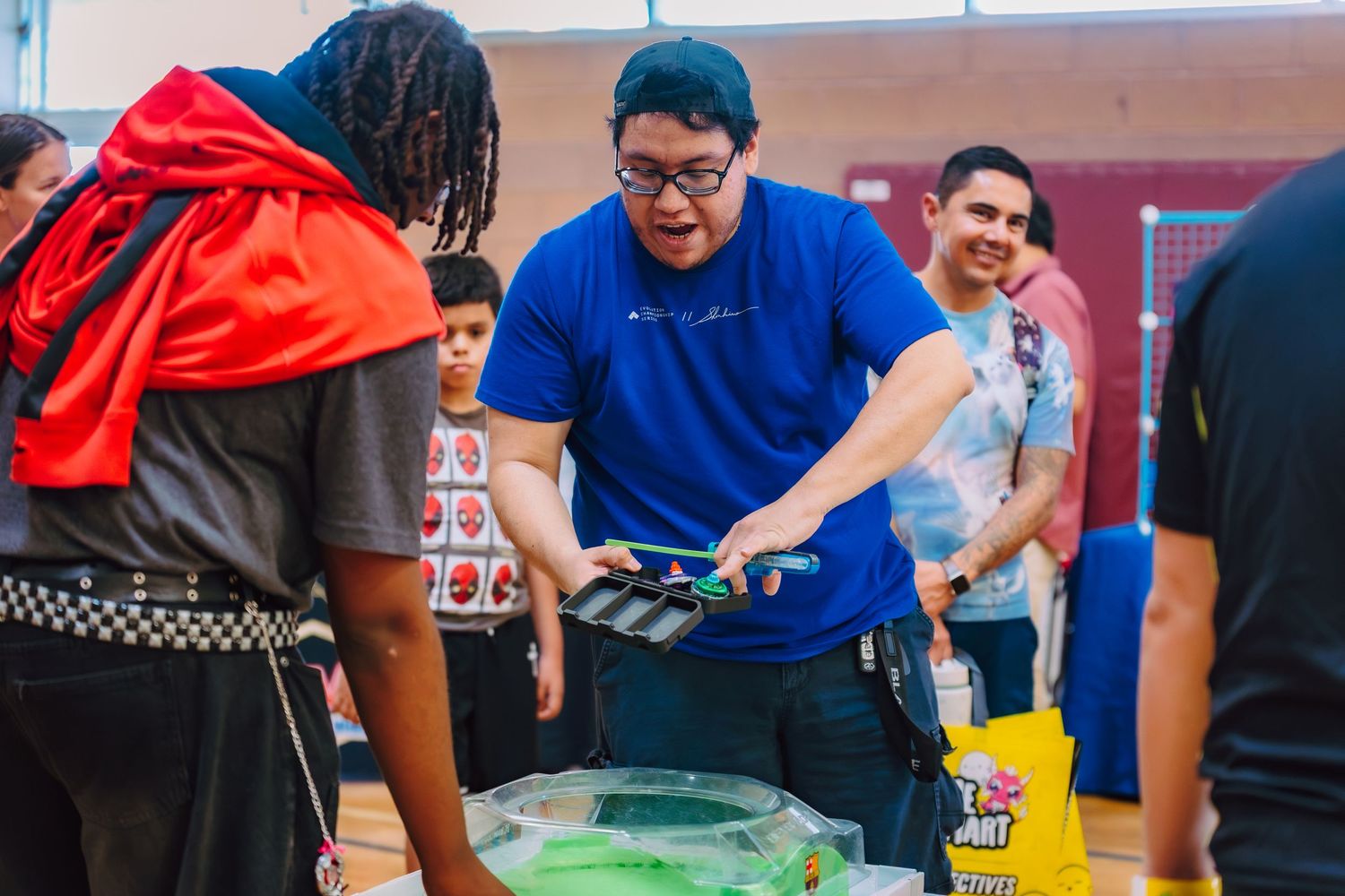 People gather around a table at an indoor community event, with a man in a blue shirt handing out items.