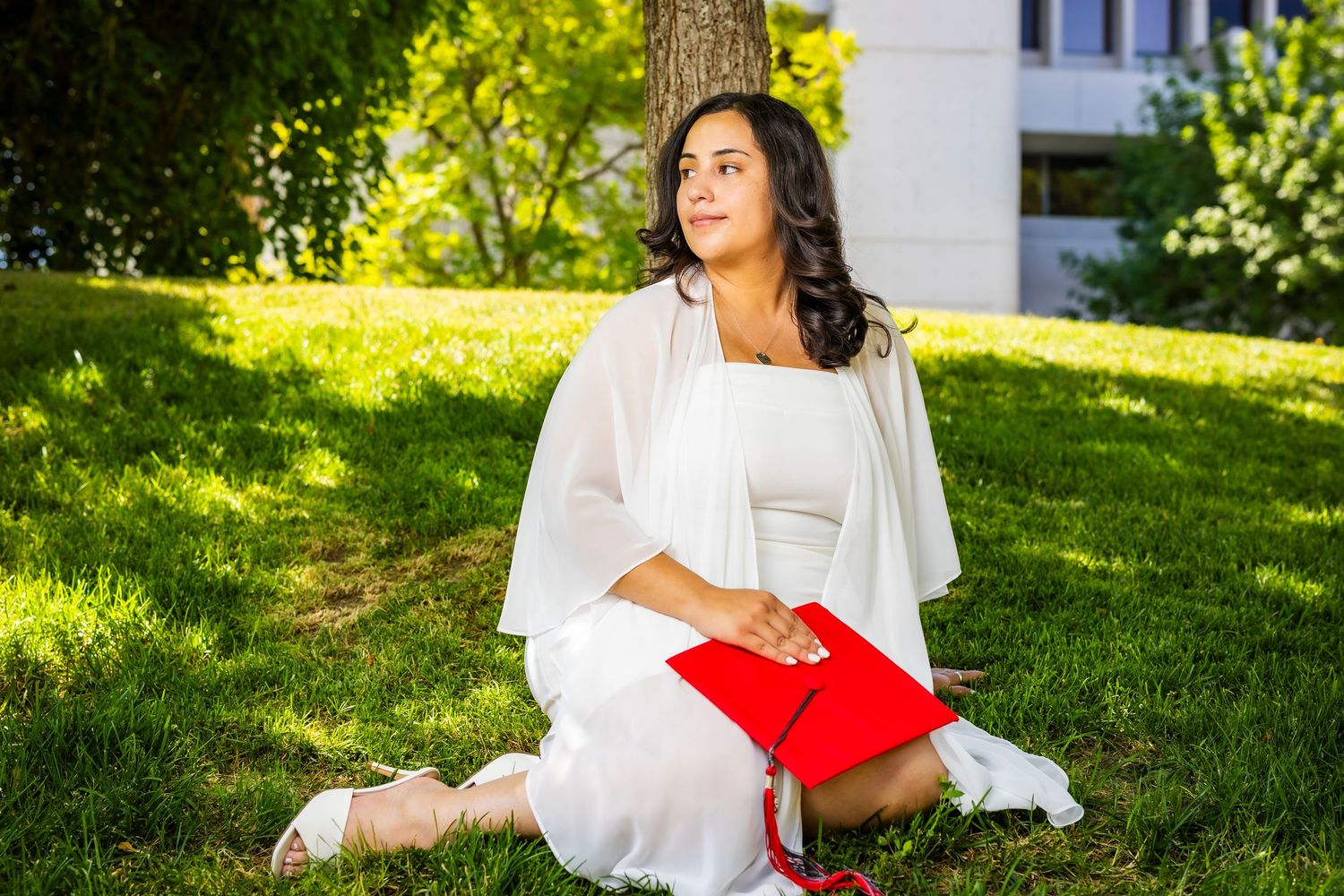 Woman in white dress holding red graduation cap sits on grass leaning against tree outdoors.