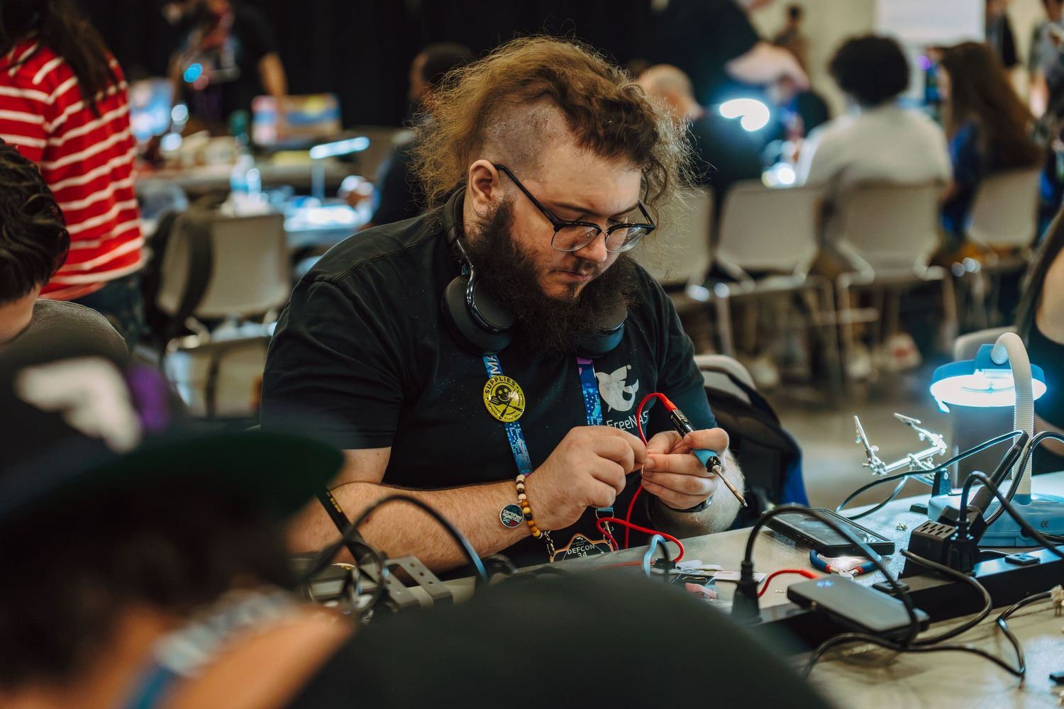 A focused technician with long hair and glasses soldering electronics at a crowded maker event or hackathon.