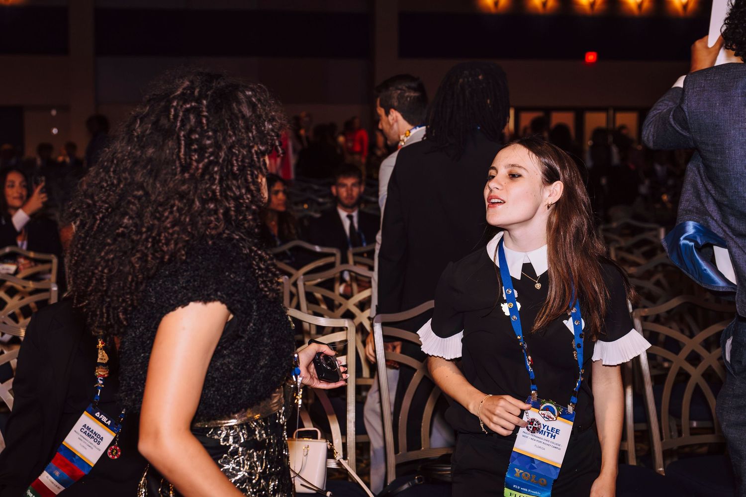Two women networking at a formal evening event, wearing lanyards and badges in a dimly lit conference venue.