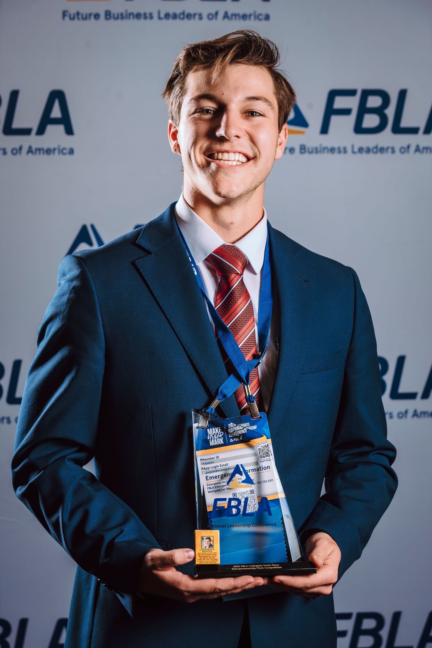 Young man in blue suit holding FBLA award trophy, smiling at camera in front of Future Business Leaders of America backdrop.