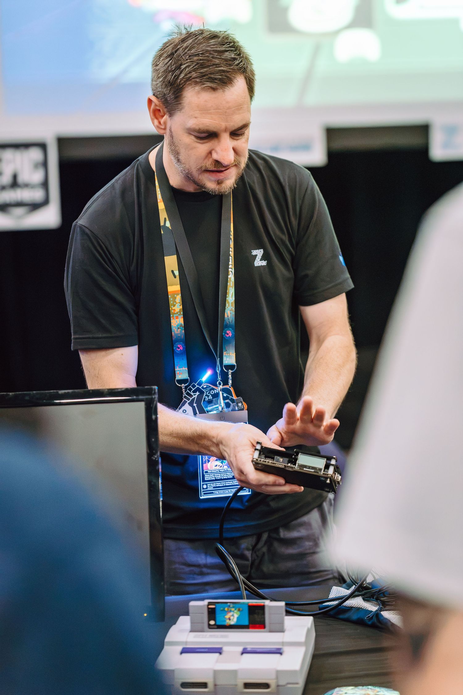 Man in black t-shirt demonstrating electronics hardware at a gaming convention booth with SNES console on table.
