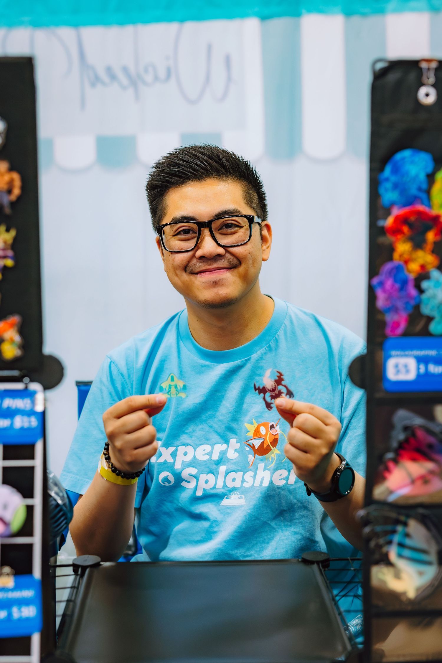 Smiling young man in blue Expert Splasher t-shirt holding small figurine at a colorful gaming convention booth.