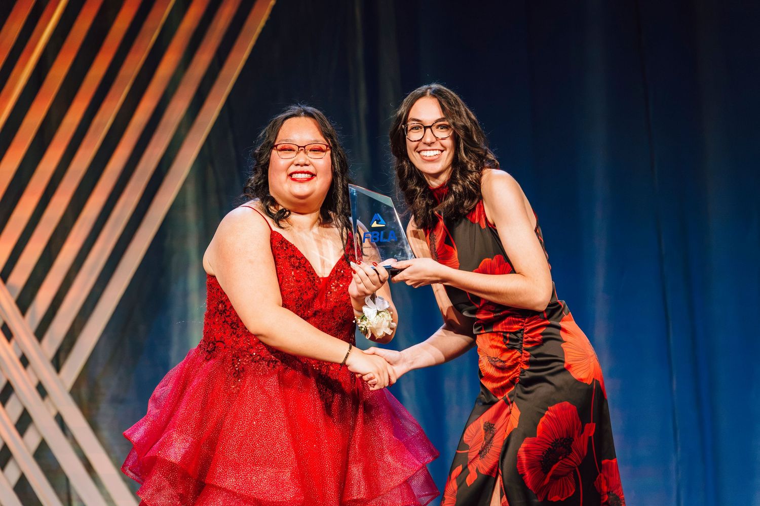 Two women in red dresses share an award on stage at a formal ceremony event.