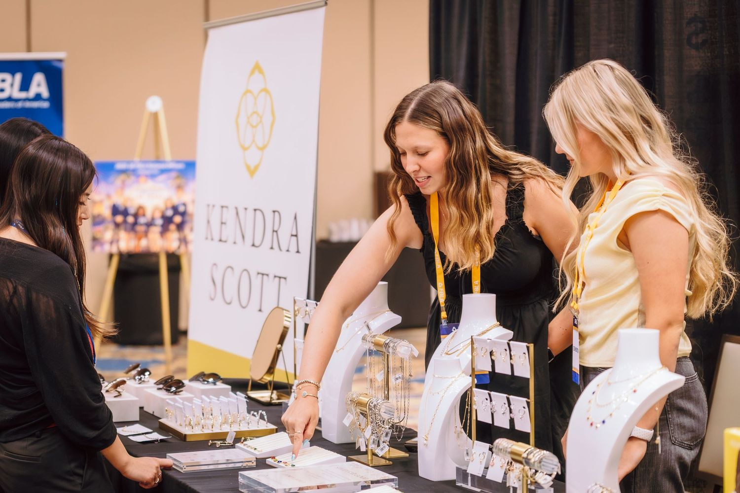 Two women browsing jewelry at a Kendra Scott display booth at a business conference expo.