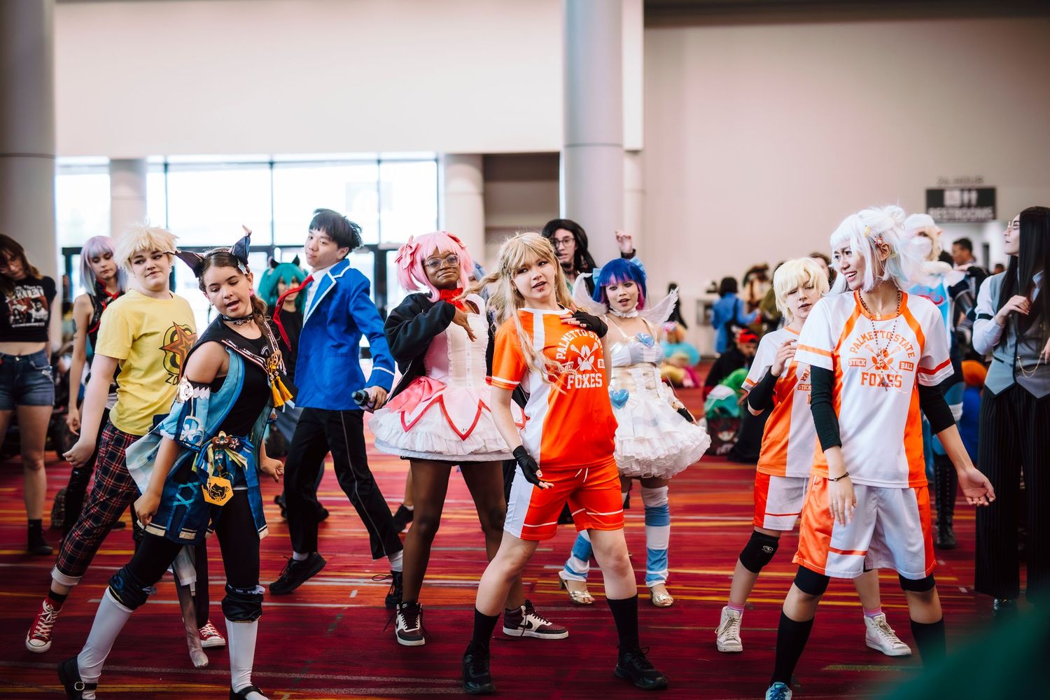 Group of anime cosplayers in colorful costumes performing a dance routine at a convention hall.