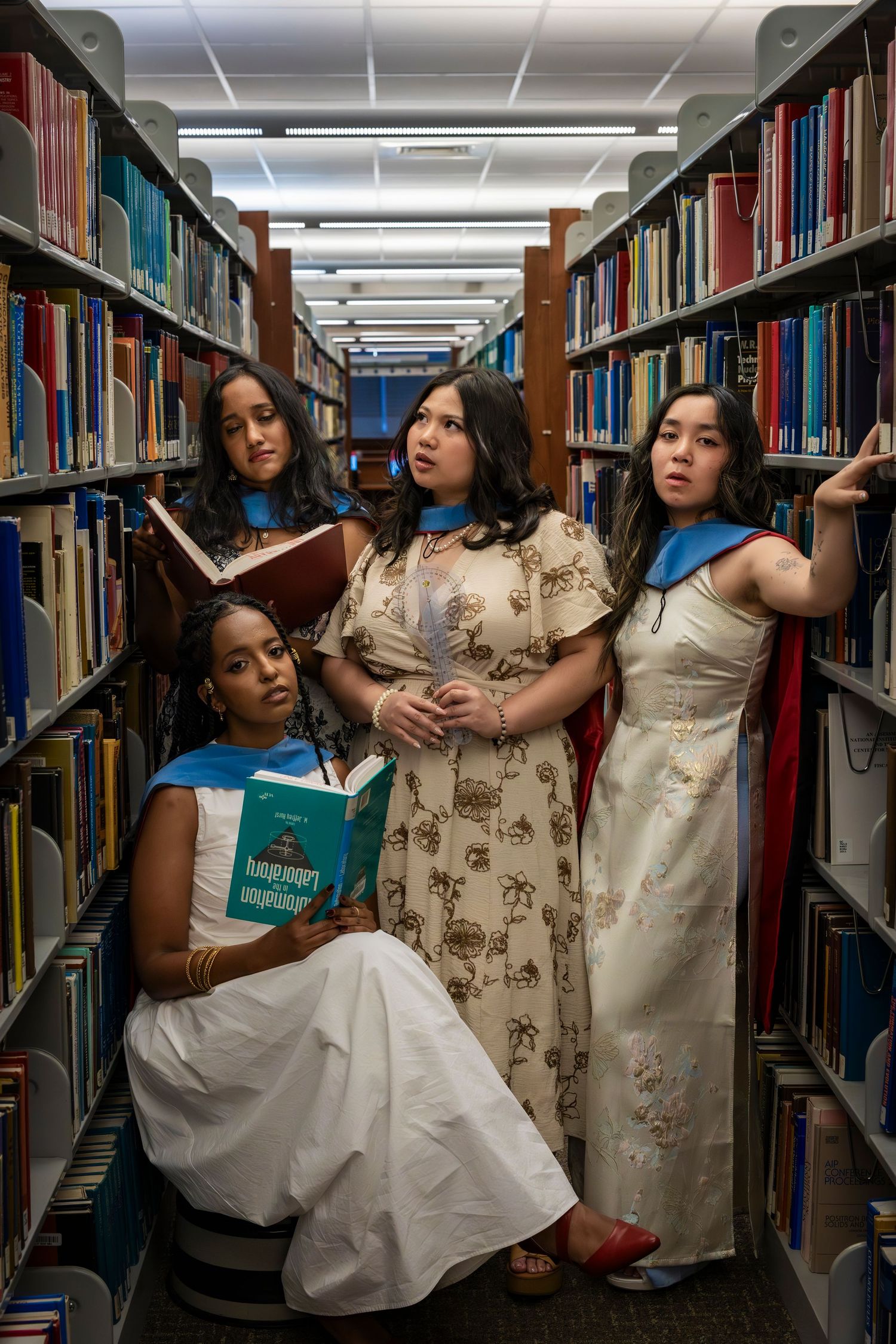 Four diverse women in elegant dresses pose dramatically among library bookshelves, holding books in a scholarly setting.