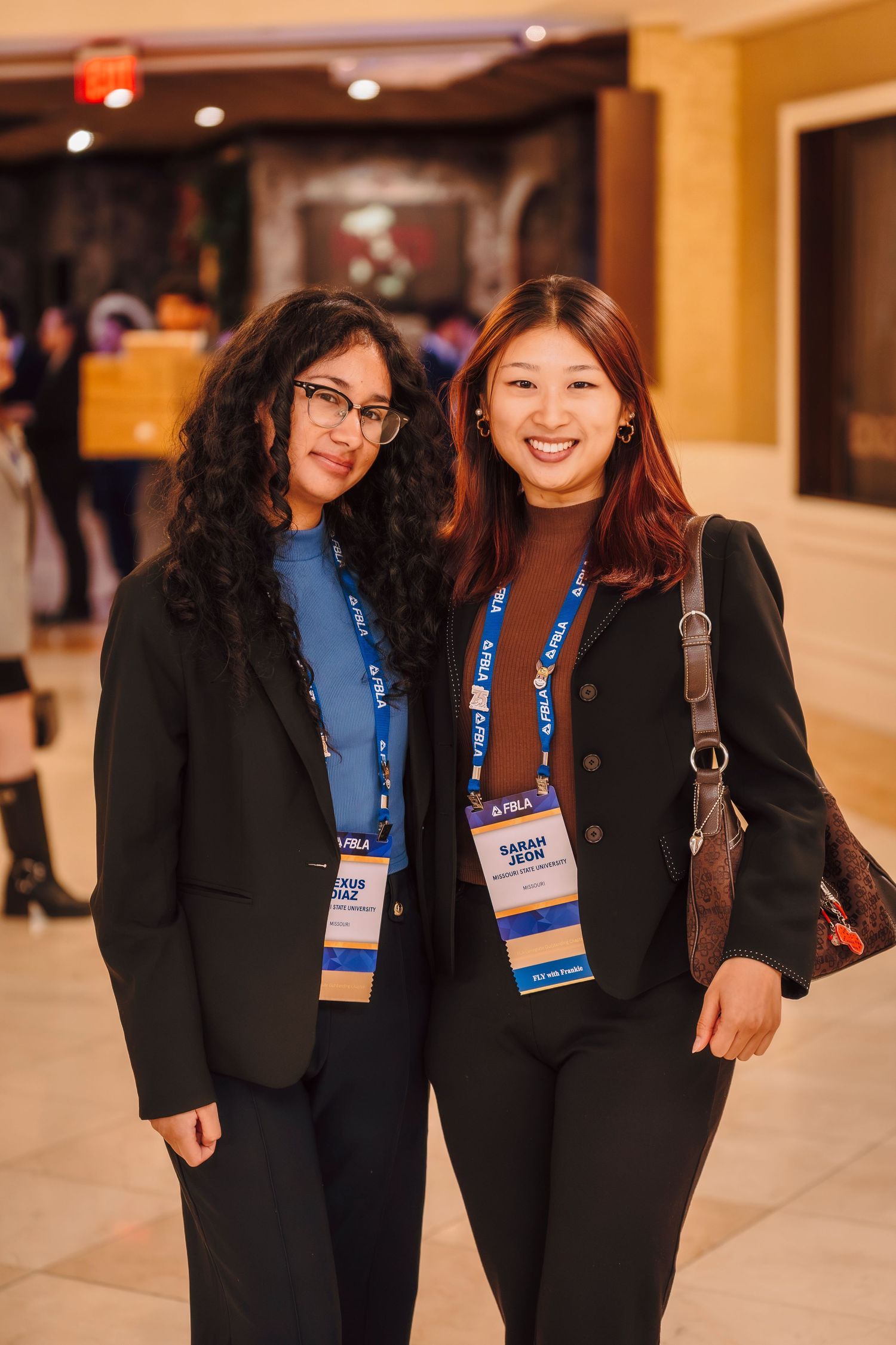 Two women wearing conference lanyards and black blazers smile together at a professional event.