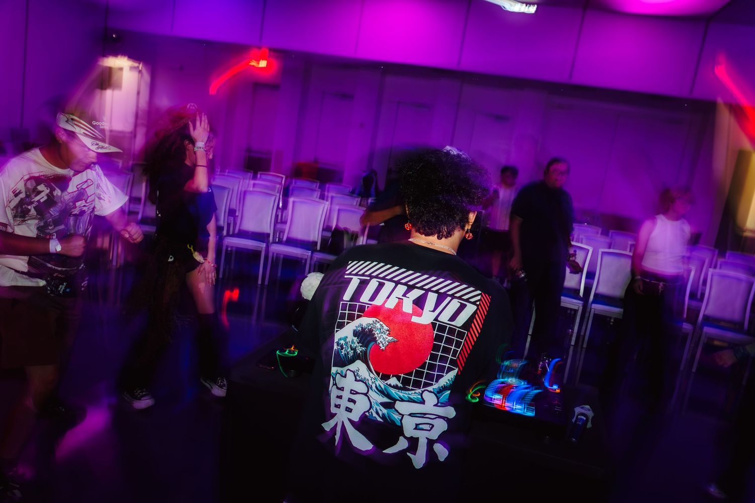 People dancing at a neon-lit party, featuring a Tokyo graphic bomber jacket under purple and pink lights.
