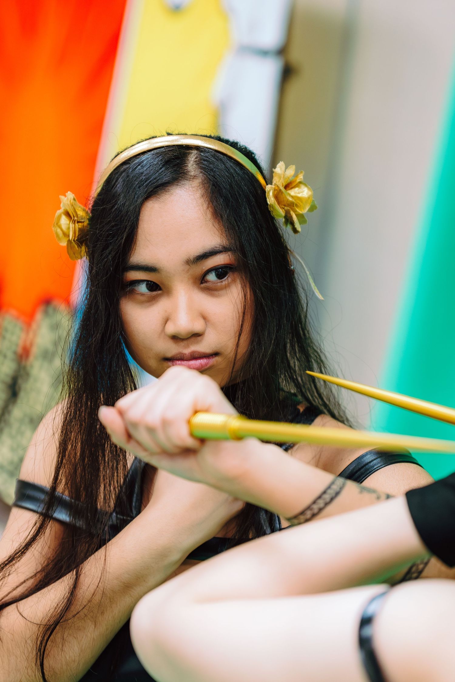Young Asian woman in cosplay costume with gold floral headpiece, holding golden arrows in a dramatic pose.