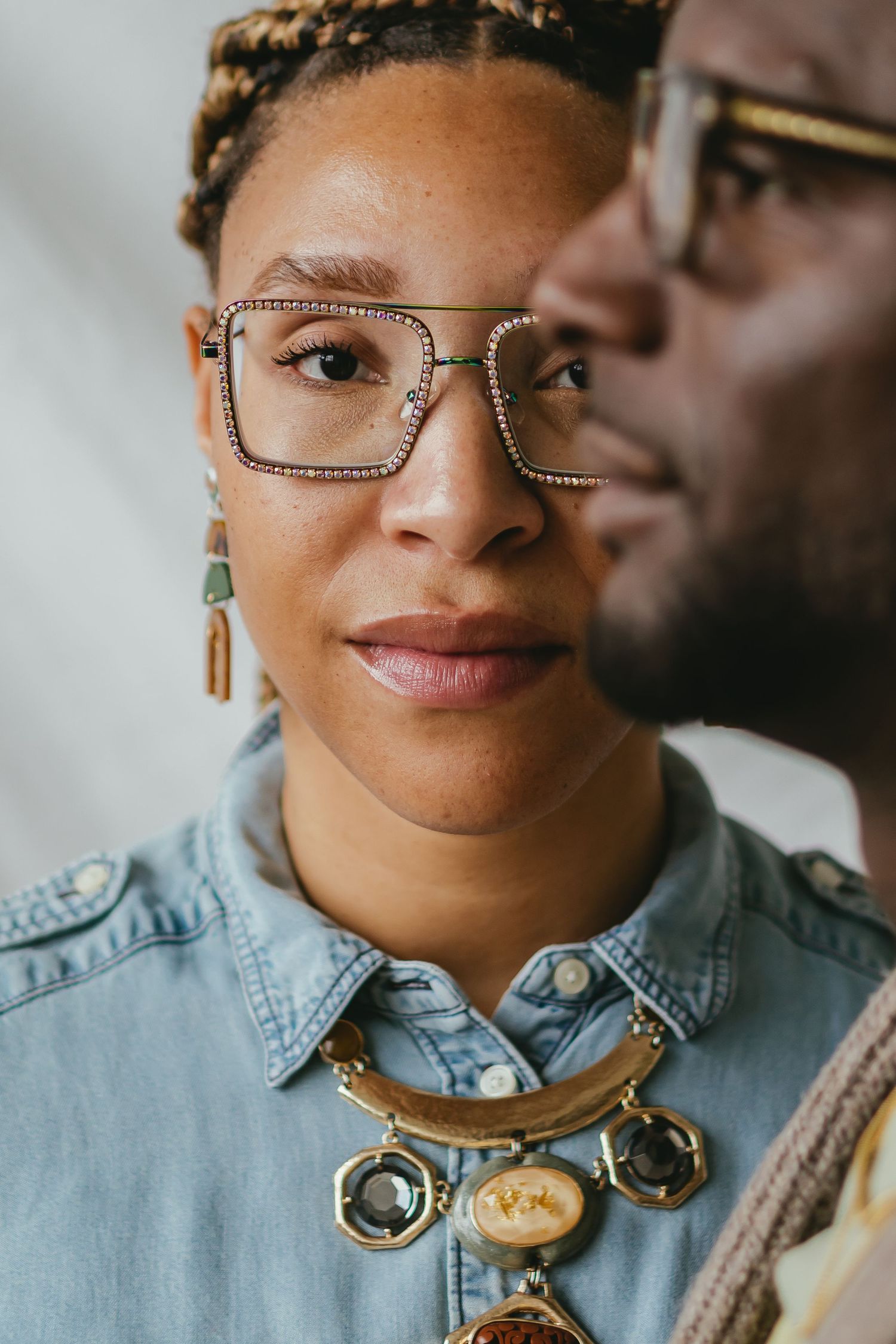 A stylish Black woman wearing rhinestone glasses and a gold statement necklace poses with a man in glasses.