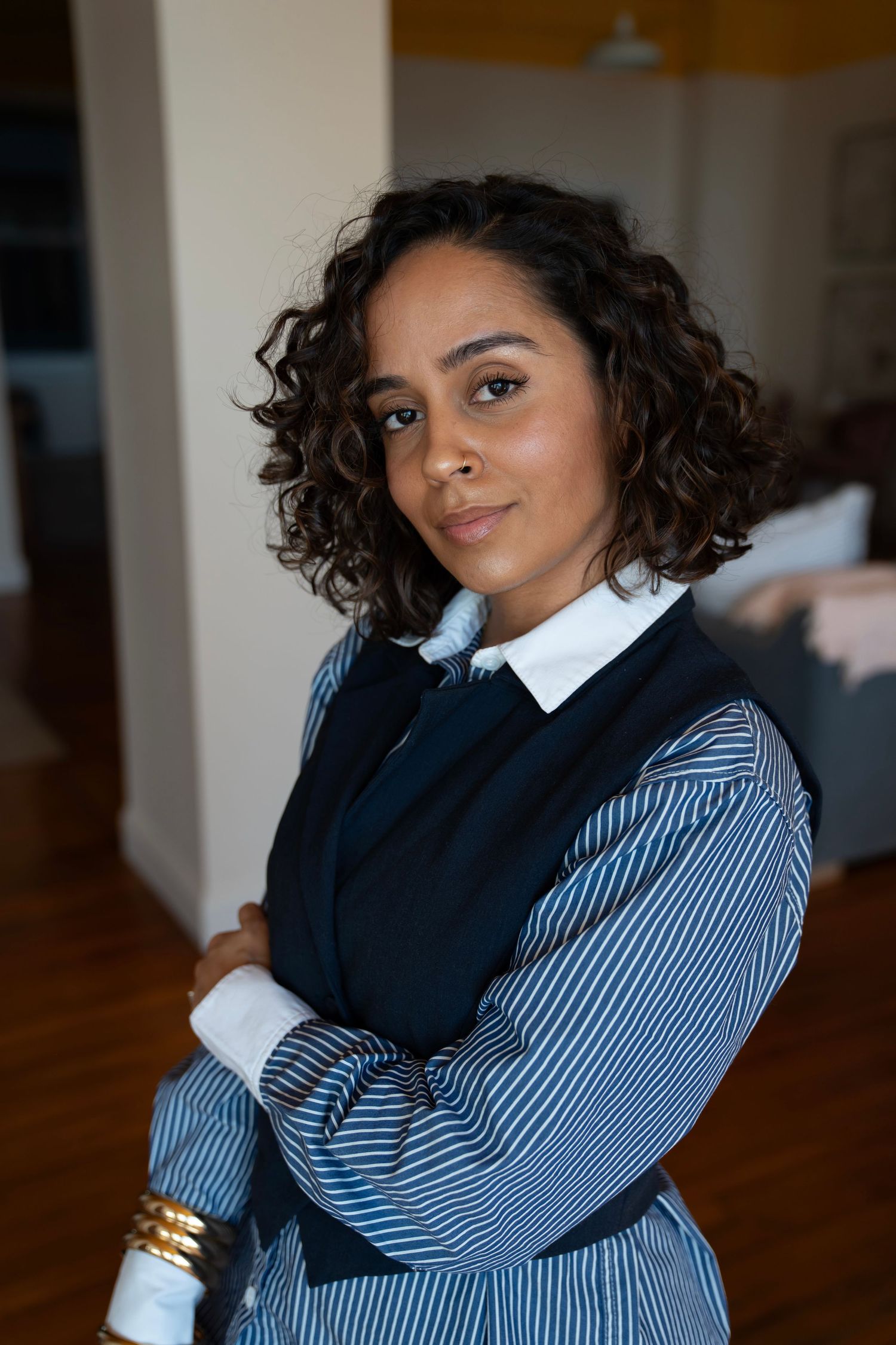 Professional woman with curly hair, arms crossed, wearing striped shirt and navy vest, gold bracelet, indoors.