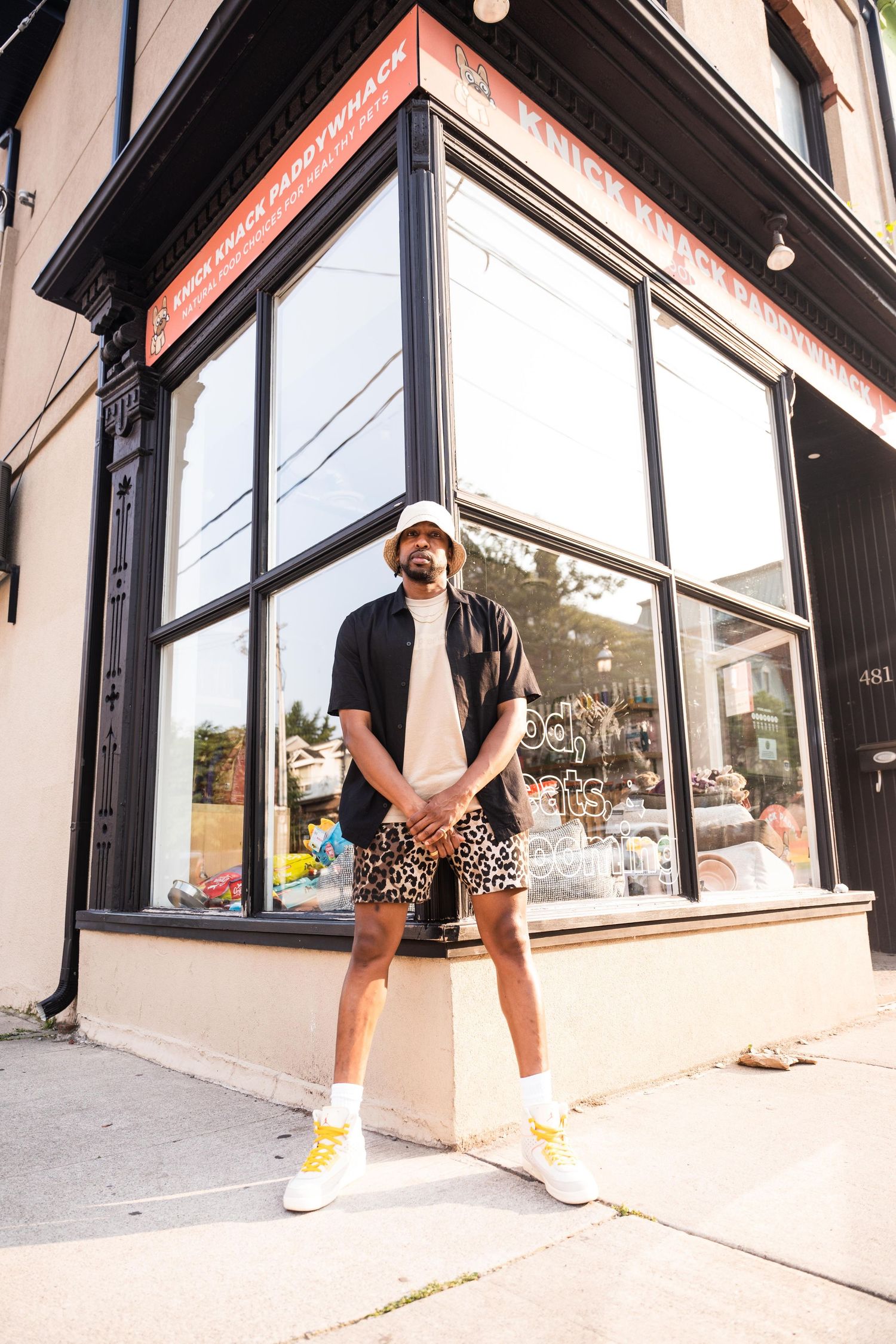 A man wearing shorts and a hat stands in front of a store, smiling and looking towards the camera.