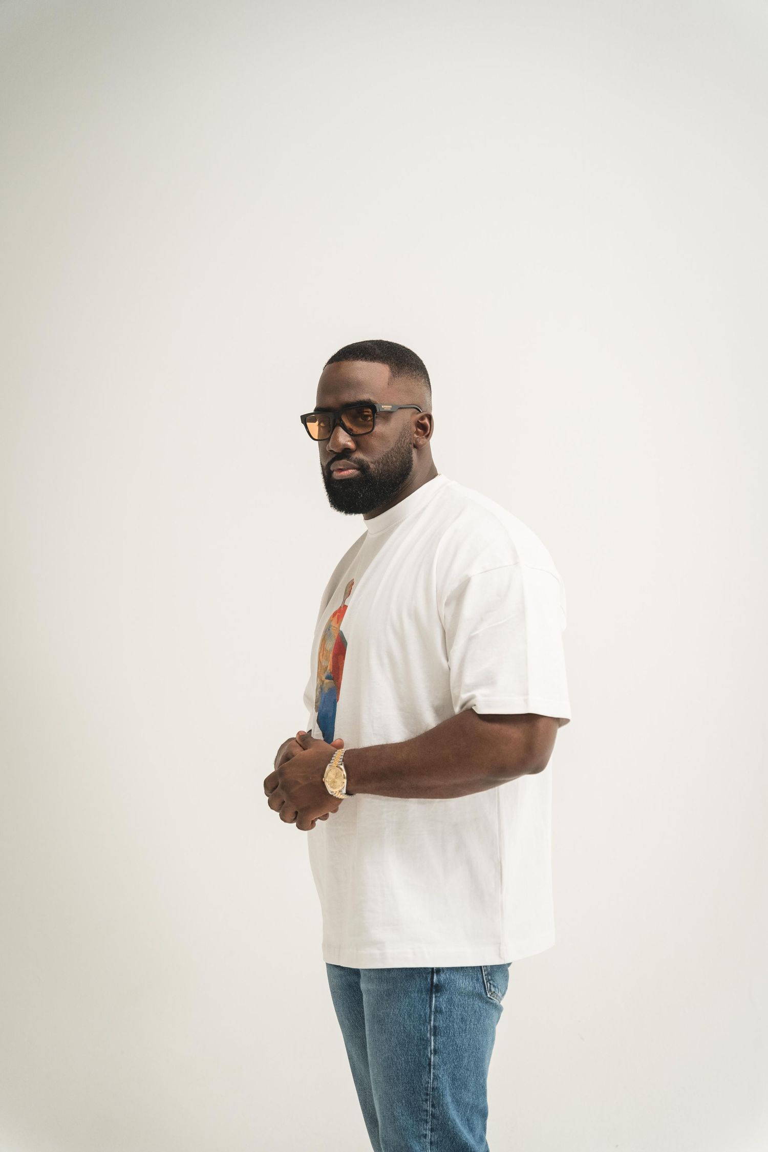 . A bearded man wearing glasses stands against a plain white wall, looking directly at the camera.
