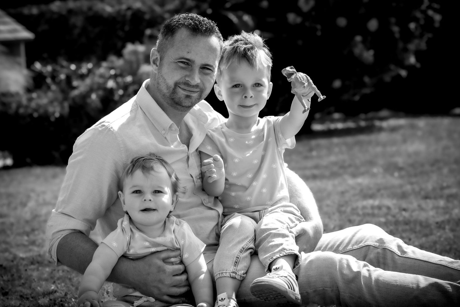 Black and white photo of a father sitting on grass with two young children, smiling outdoors in a garden setting.