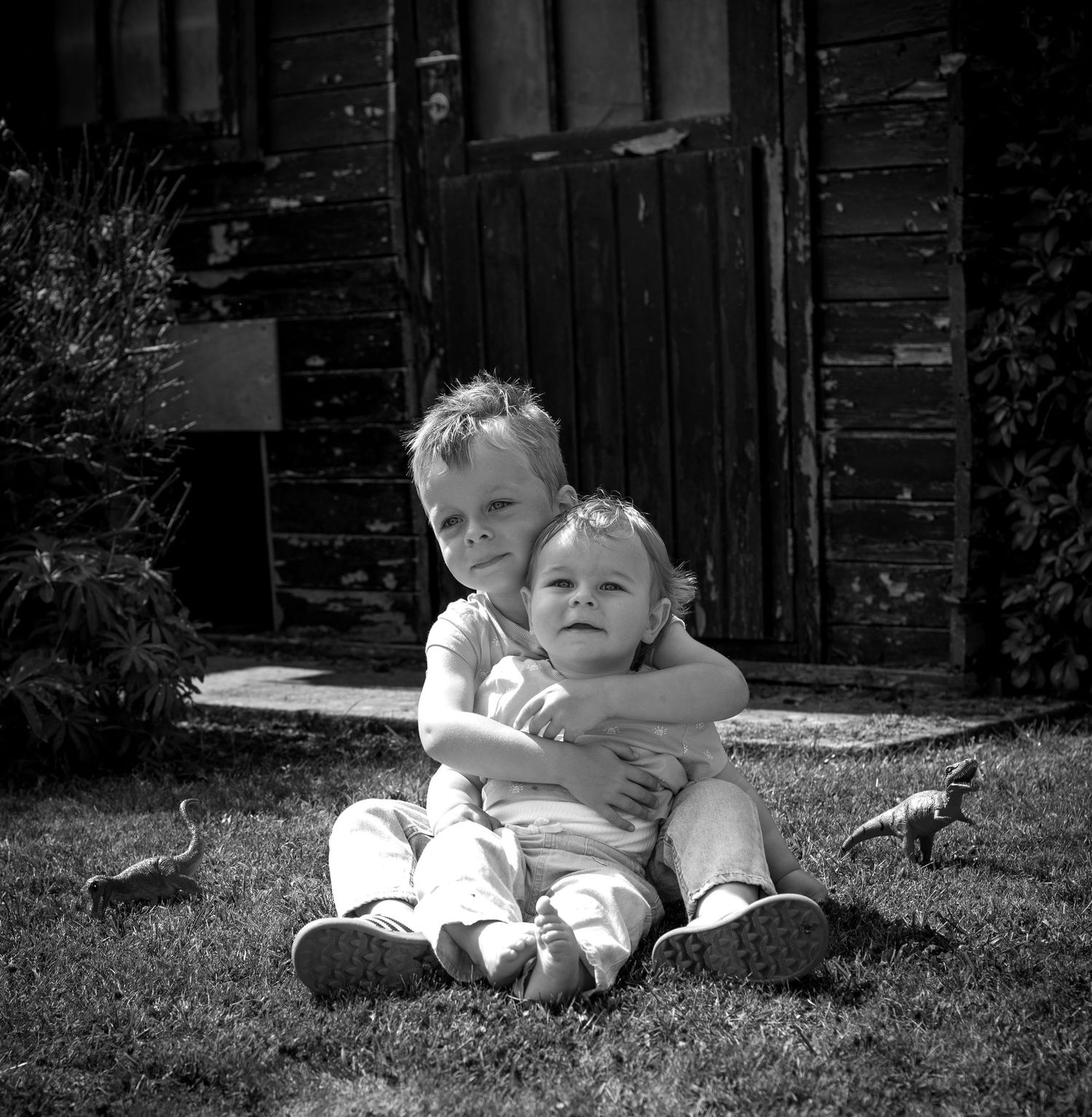 Black and white photo of two young siblings hugging on grass in front of a wooden shed.