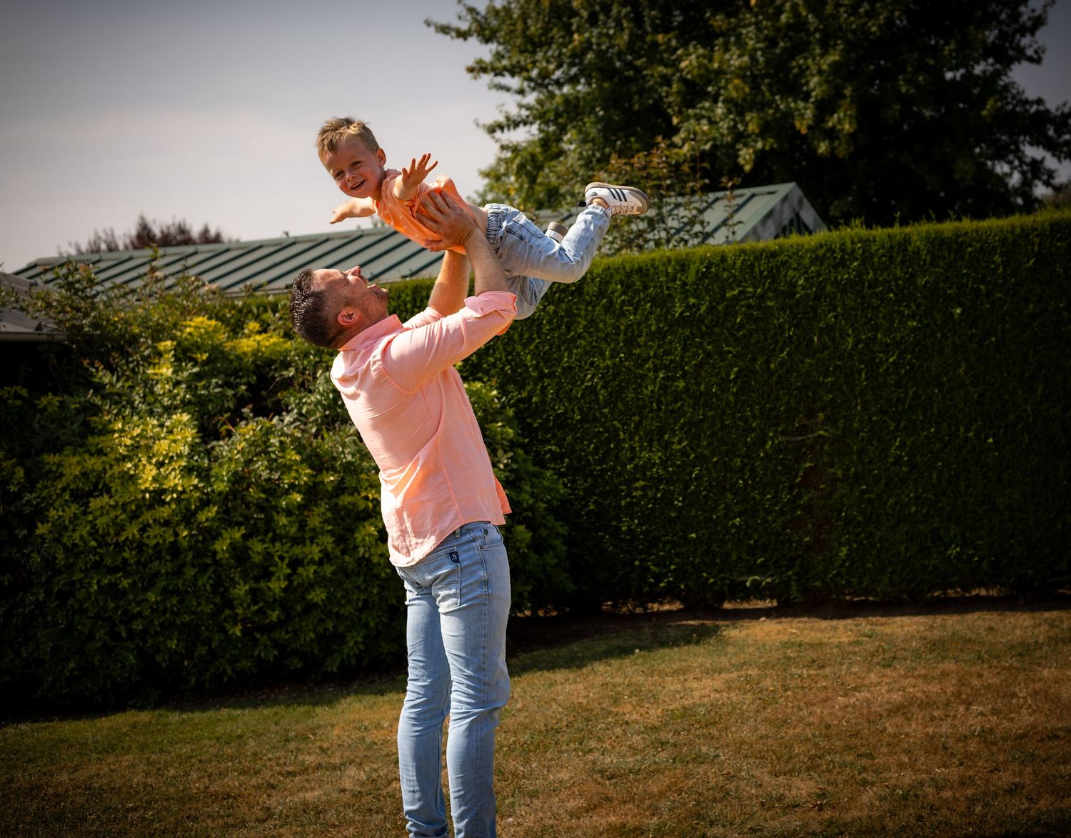 A parent joyfully lifts a child high in the air outdoors in a lush green garden on a sunny day.