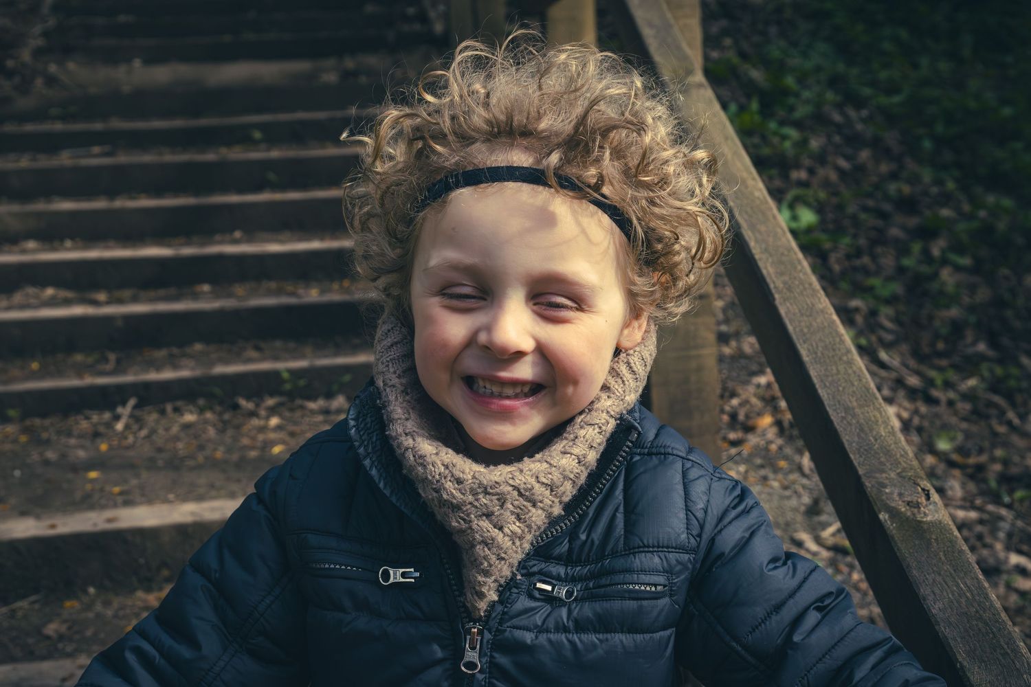 Smiling curly-haired child wearing a blue jacket and scarf headband, sitting on wooden steps outdoors.