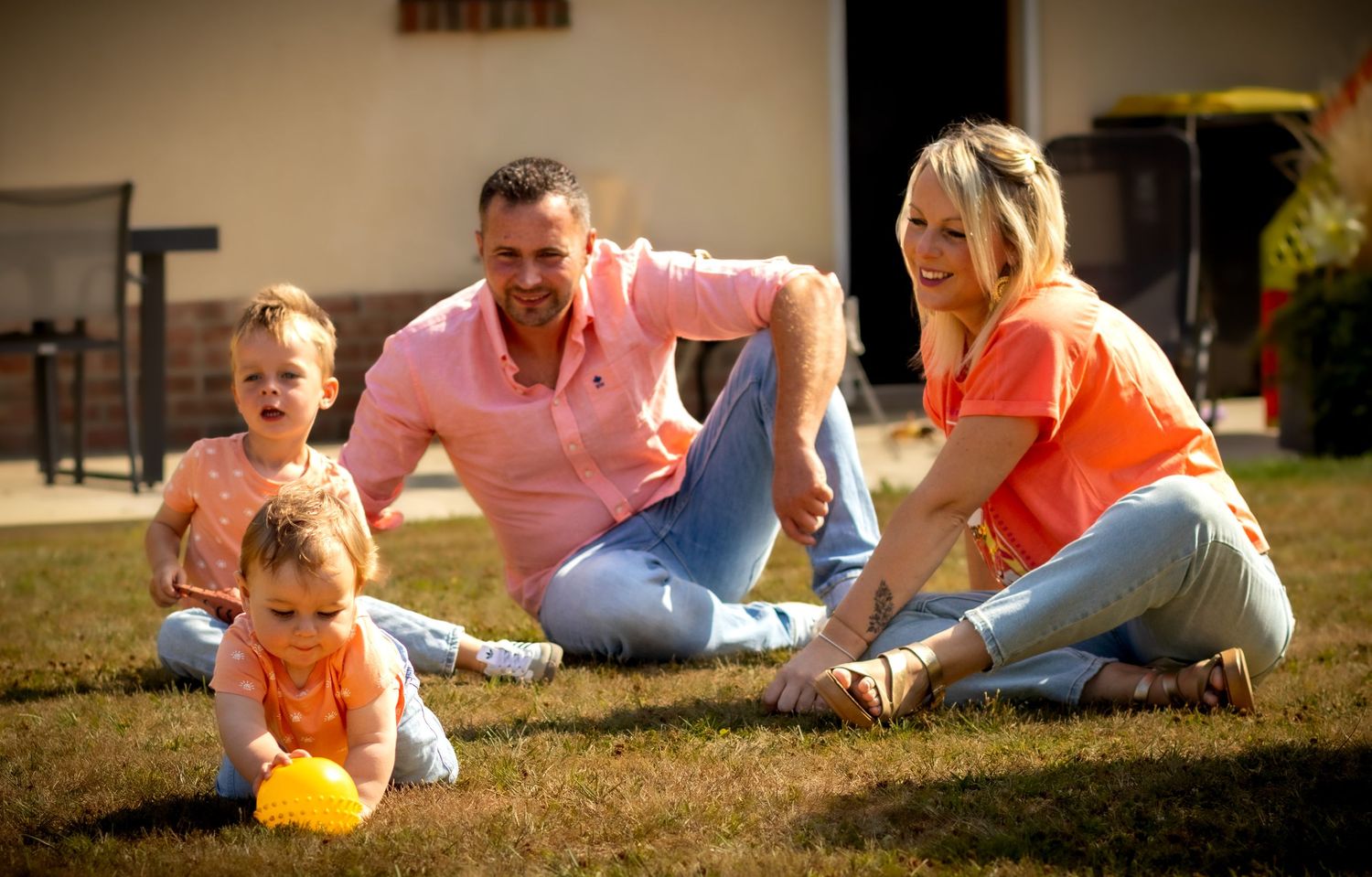 Happy family with two young children sitting on green grass in sunny backyard, parents in orange tops smiling.