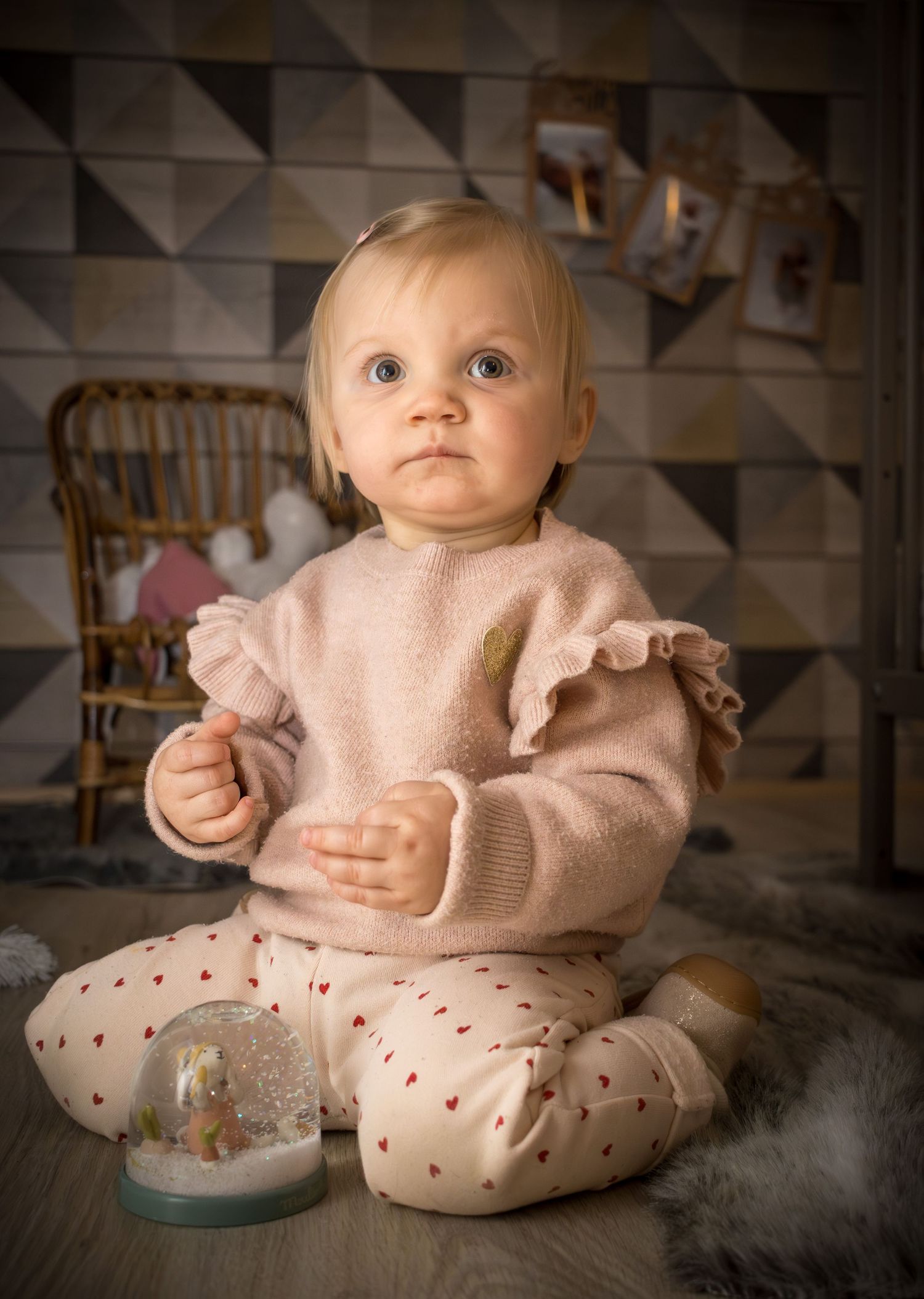 Adorable baby girl in pink ruffle sweater sitting on floor with a festive snow globe in a cozy holiday setting.