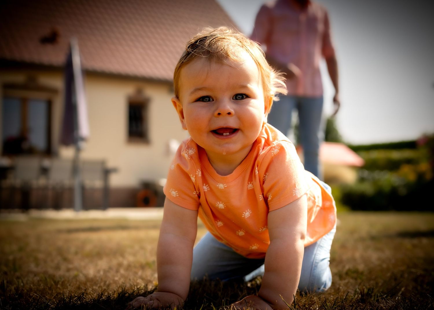 Smiling baby in orange polka dot shirt crawling on grass in sunny backyard with house in background.