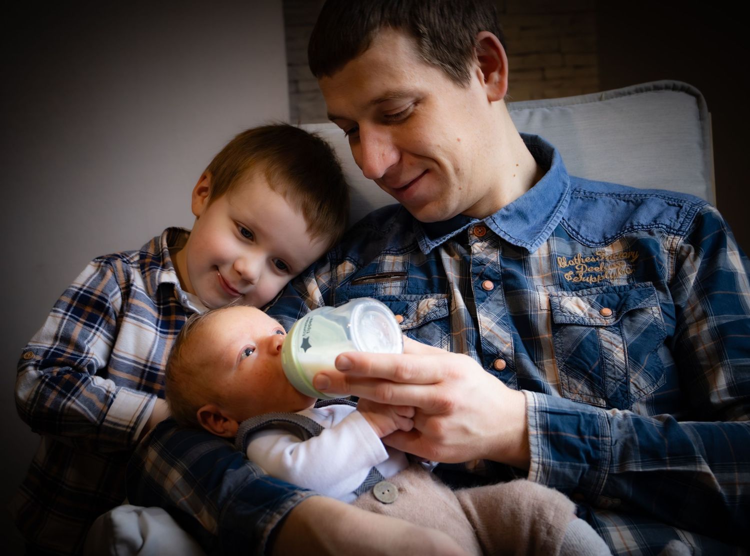 Father bottle-feeding newborn baby while young toddler son looks on, sitting together in cozy home setting.