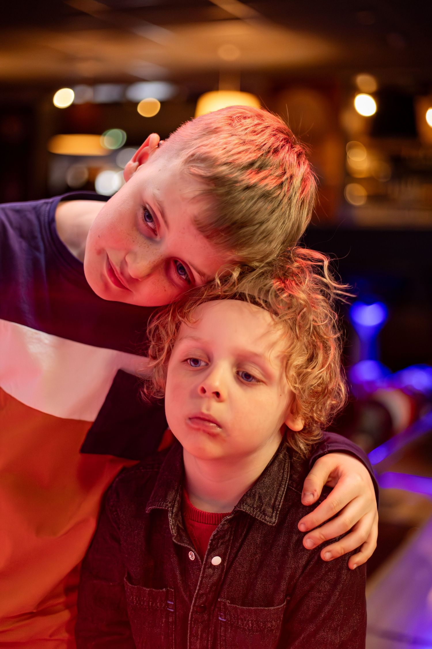 Two young boys posing together under colorful lights, older boy resting head on younger curly-haired boy's head.