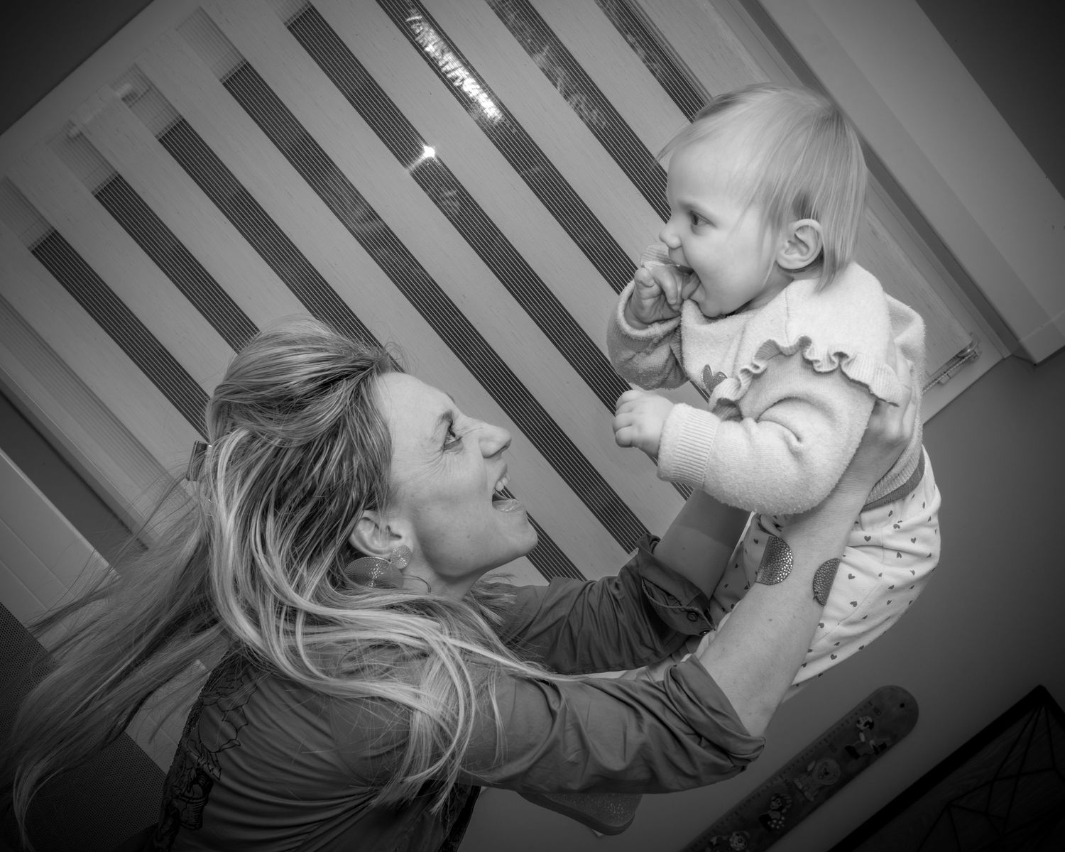 Black and white photo of a mother lovingly lifting her smiling baby up in the air near a wooden crib.