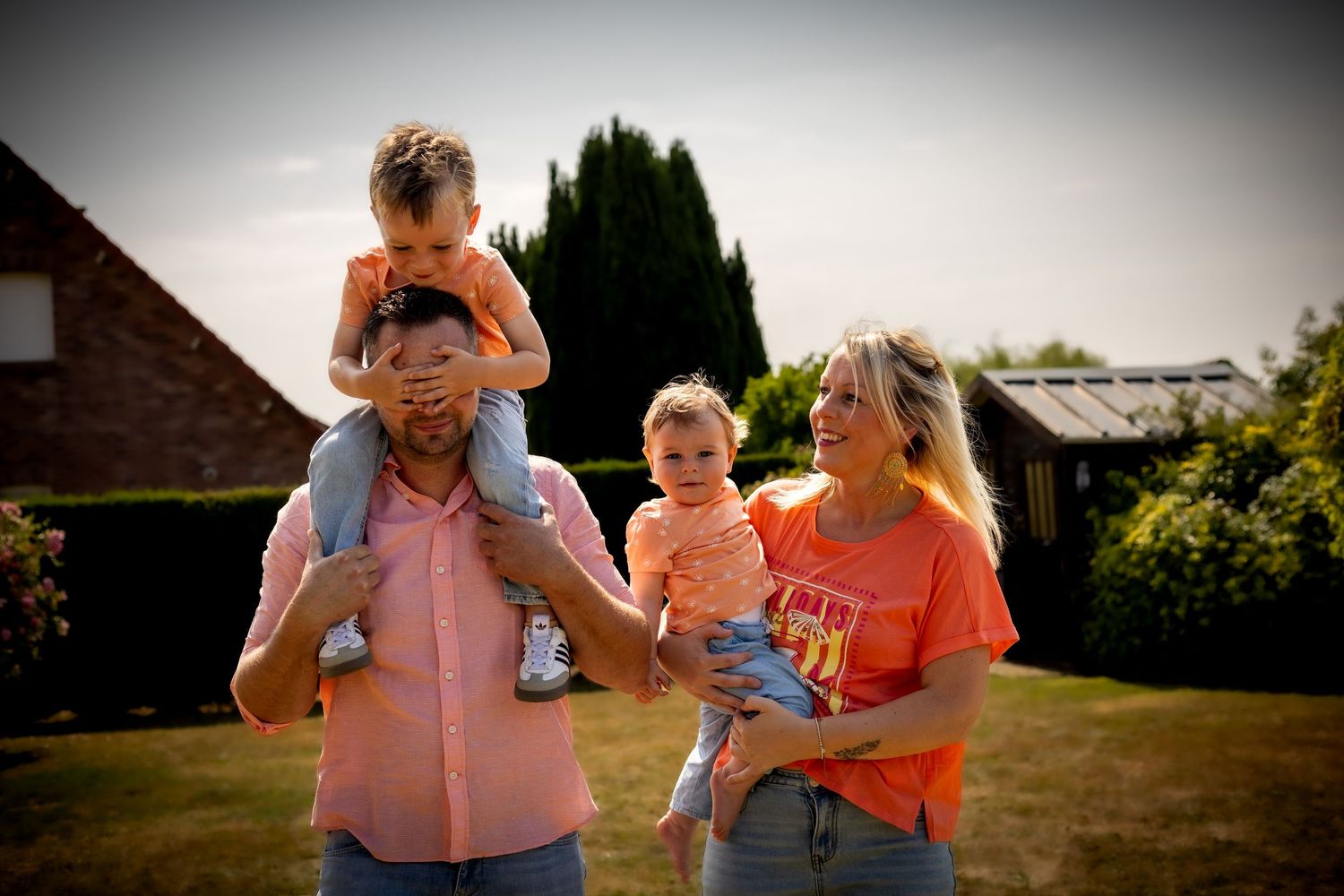 Family of four posing outdoors, father carrying toddler on shoulders while mother holds baby, all wearing orange shirts.